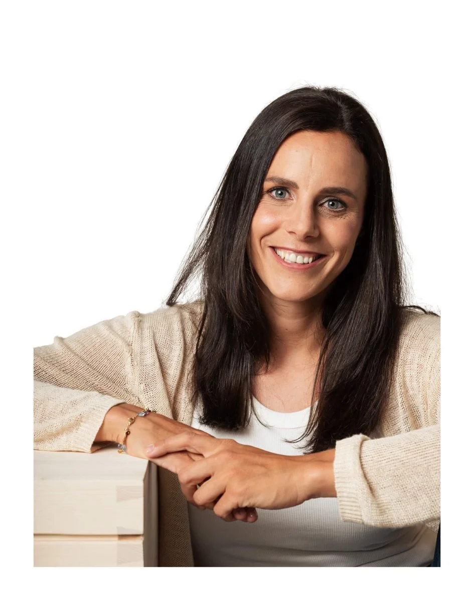 Lächelnde junge Frau mit braunen Haaren, die auf einem Tisch ruht und eine Hand auf die andere legt, vor weißem Hintergrund.