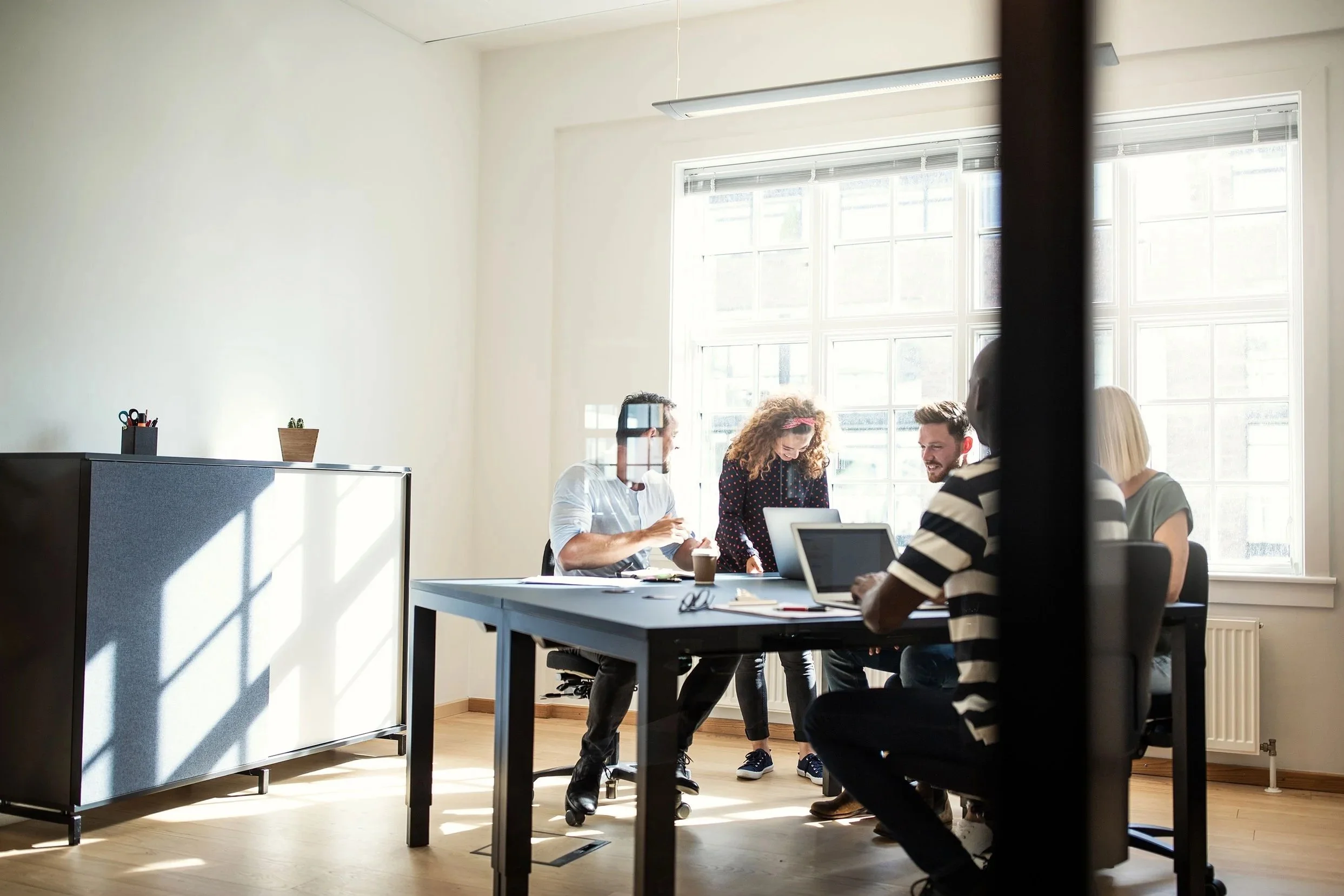 A group of five people gathered around a table in a bright meeting room, engaged in a discussion, with laptops, notebooks, and coffee cups on the table.