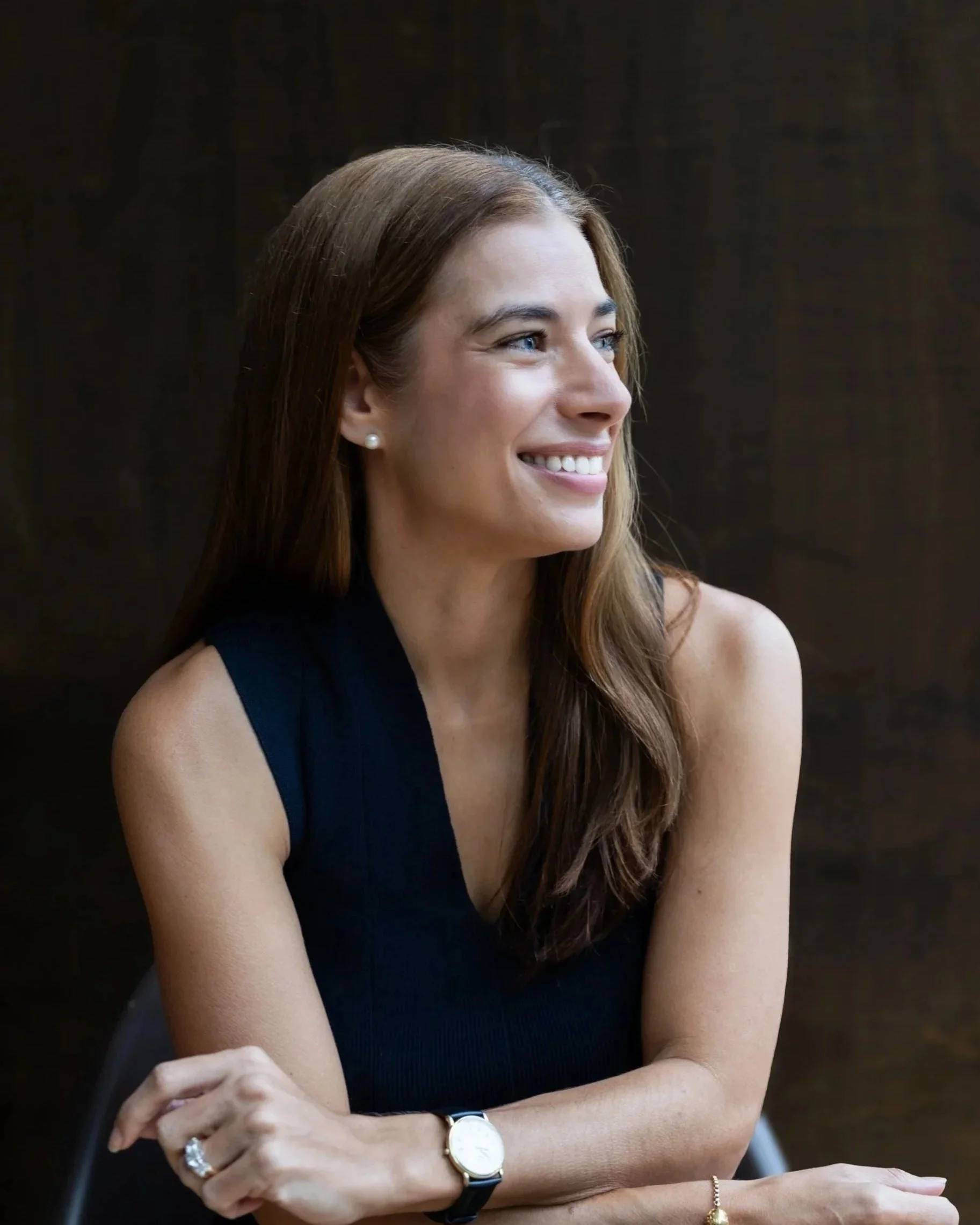 Pippa Woodhead. A woman with long, wavy auburn hair, wearing a black sleeveless top, a pearl earring, a watch, a ring, and a bracelet, sitting with her arms crossed, smiling and looking to her right against a dark background.