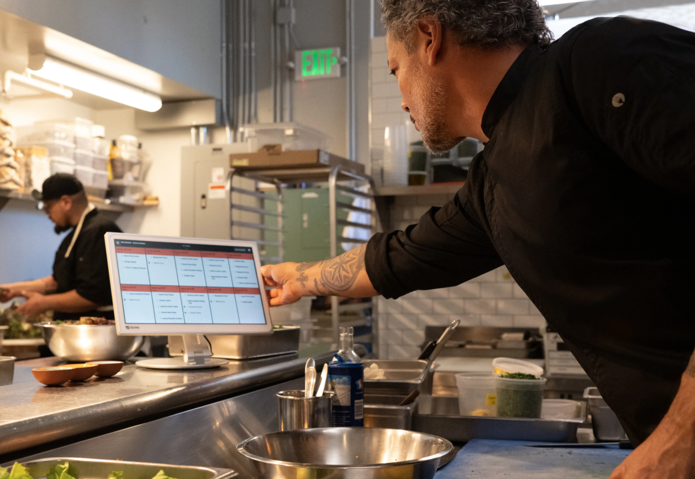 A chef in a black uniform working in a commercial kitchen, pointing at a digital menu or schedule displayed on a monitor.