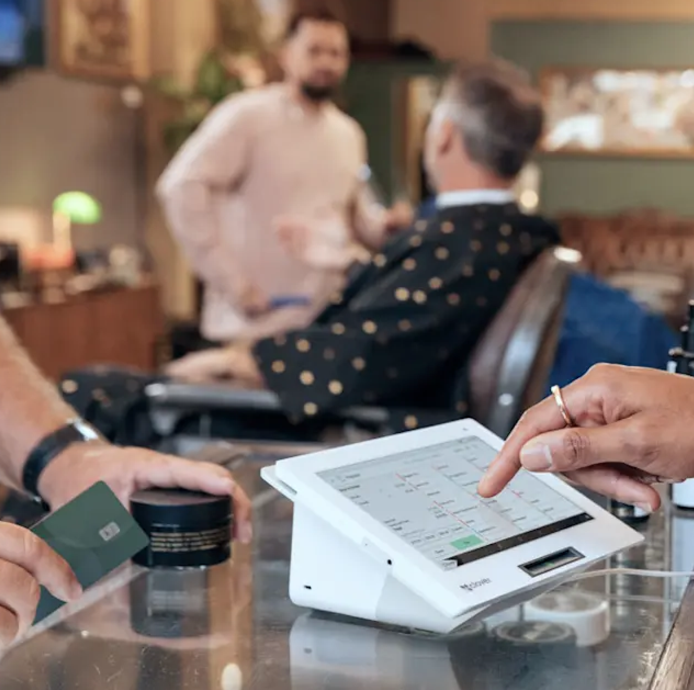 A person is using a tablet with a point-of-sale system on a counter in a cafe, with a person holding a credit card and a small jar nearby. In the background, two people are engaged in conversation.