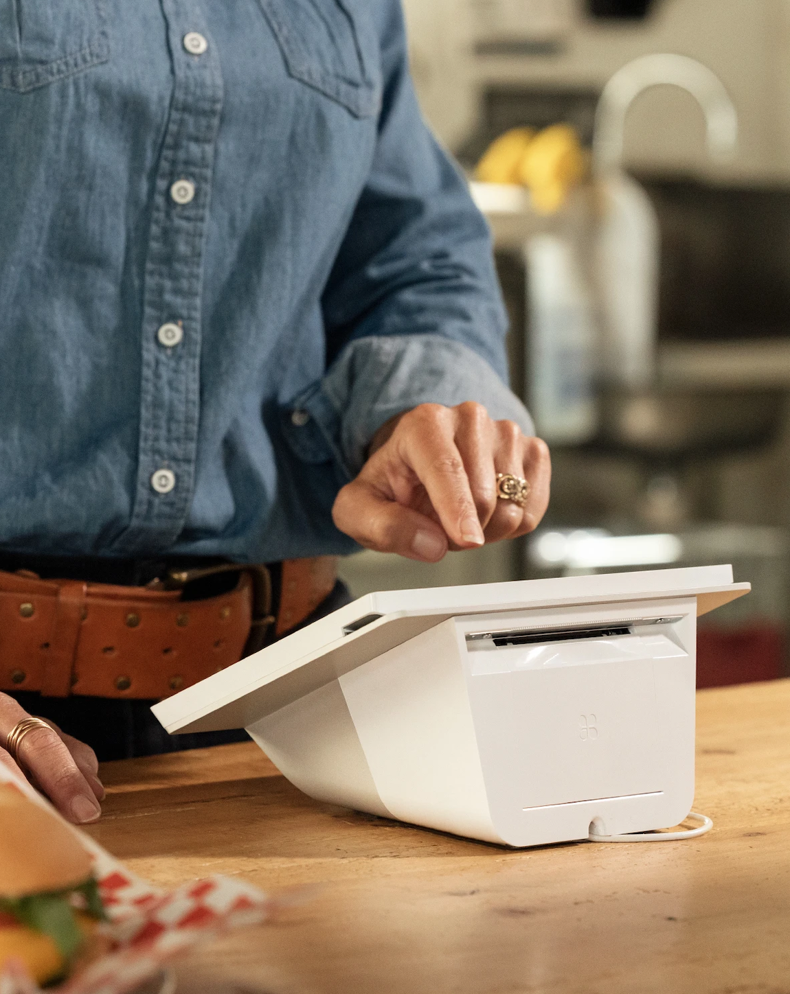 A person using a white point-of-sale terminal at a wooden counter in a kitchen setting.