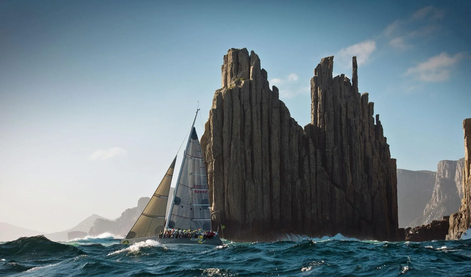 A sailboat racing in choppy ocean waters near large rock formations or cliffs under a blue sky.