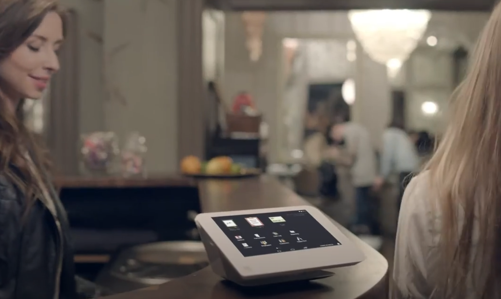 A woman at a restaurant counter using a touchscreen point-of-sale device, with another woman partially visible on the right and blurred background of the restaurant interior.