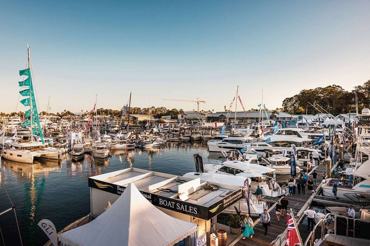 A busy marina filled with yachts and boats docked along the pier, with people walking around and boat sales booths in the foreground during sunset.