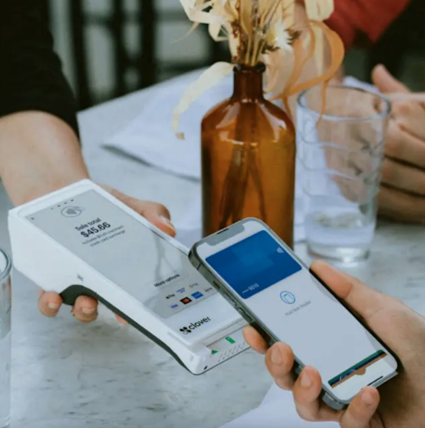 Person making a contactless payment with a mobile phone at a table with glasses of water and a brown vase with dried flowers.
