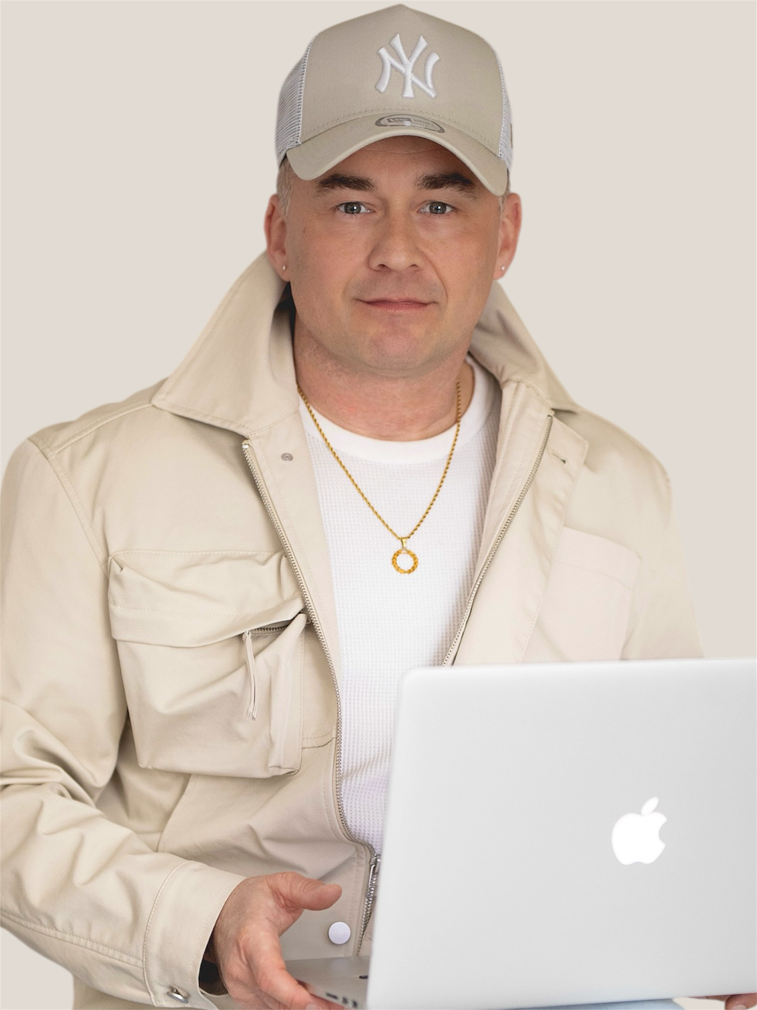 Ein Mann trägt einen beige Baseballcap mit NY-Logo, beige Jacke, weiße Shirt, Goldkette, steht vor einem MacBook und schaut in die Kamera.