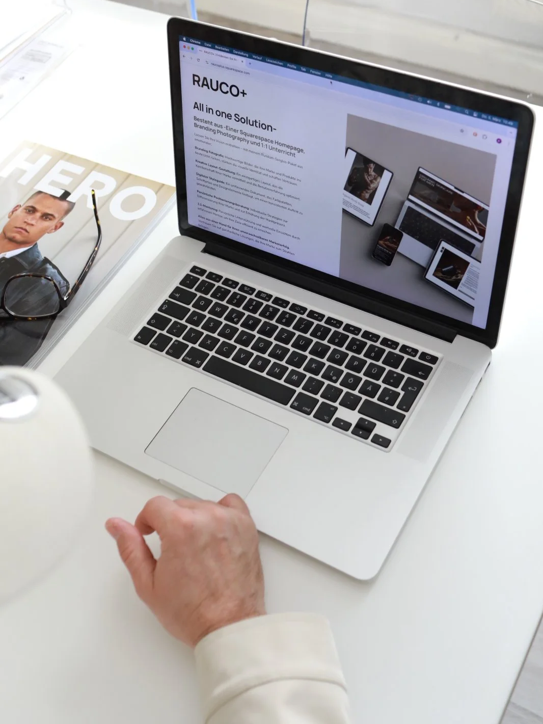 Laptop auf einem weißen Tisch, zeigt eine Webseite mit dem Titel "RAUCO+" und einer Produktbeschreibung. Daneben liegen eine Sonnenbrille, eine Zeitschrift mit einem Porträt eines Mannes und einer Brille, und eine weiße Vase.