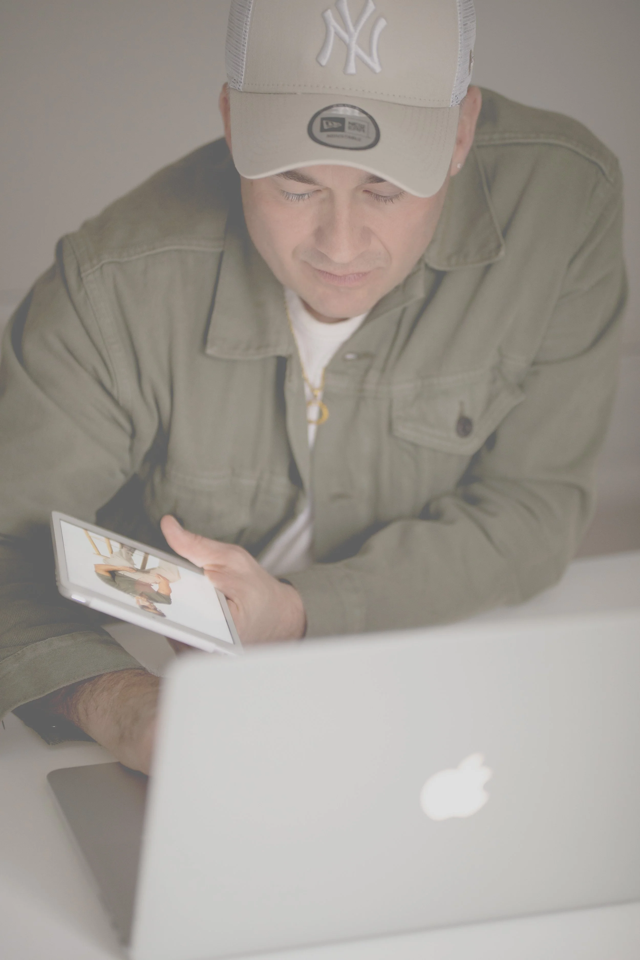 Junge Person mit Cap, Kette und Ohrpiercing, benutzt Tablet und Laptop, sitzt an einem Tisch, neutrale Hintergründe.