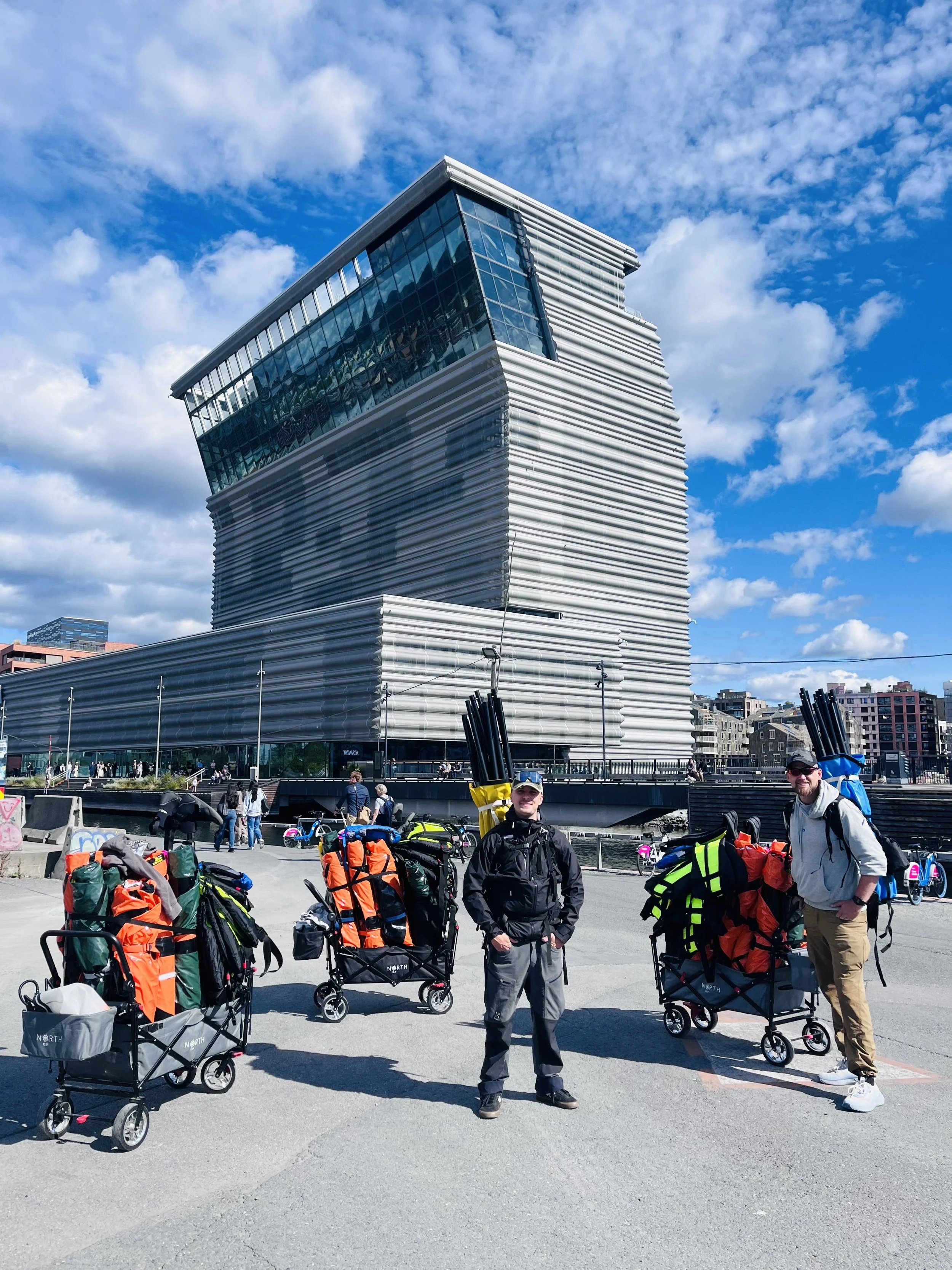 Two men standing outdoors in front of a modern building with a unique architectural design, surrounded by multiple luggage carts loaded with colorful backpacks and gear, under a partly cloudy sky.