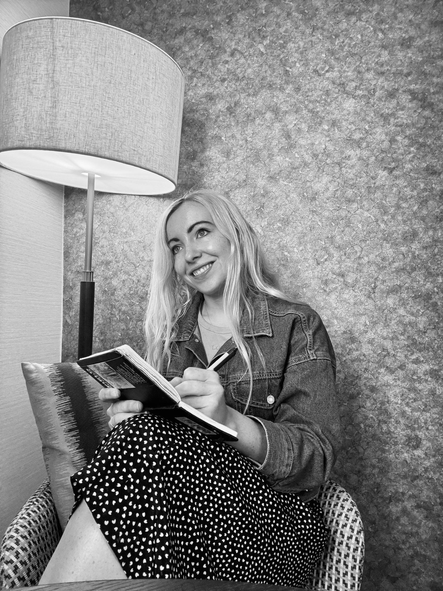 A young woman with long blonde hair sitting in a patterned chair, smiling while holding a magazine and a pen, with a lamp and patterned wallpaper in the background.