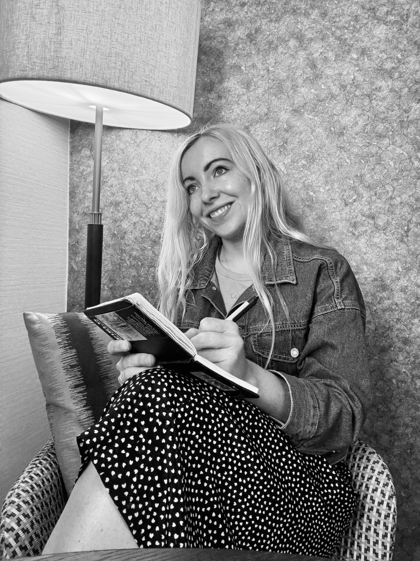 A young woman with long light hair sitting on a chair, smiling, holding a notebook and pen, near a lamp and textured wall.