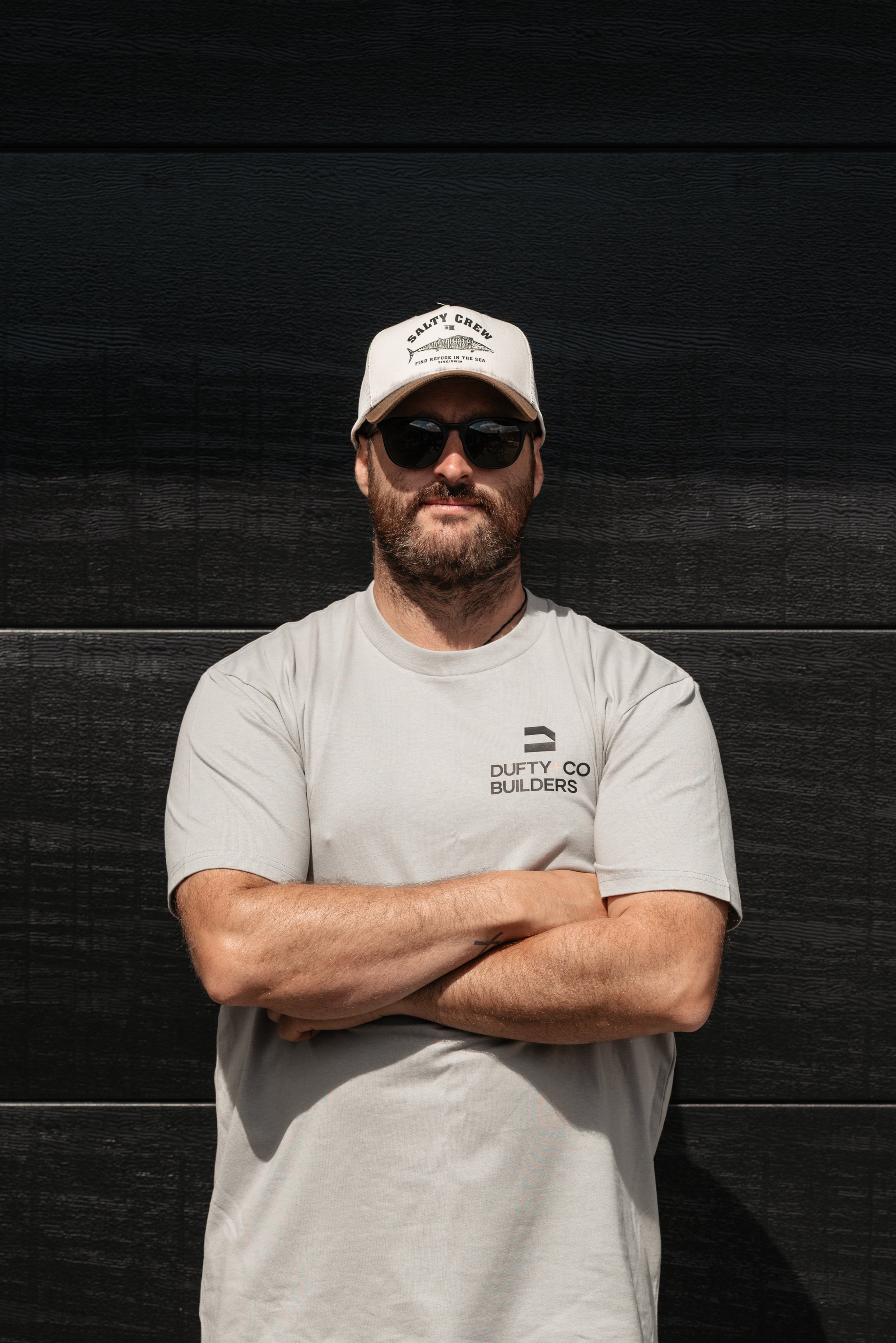A man with a beard and sunglasses standing in front of a dark wooden wall, wearing a light gray t-shirt with 'DUFTY & CO BUILDERS' on it and a white cap with 'SALTY CREW' written on it.