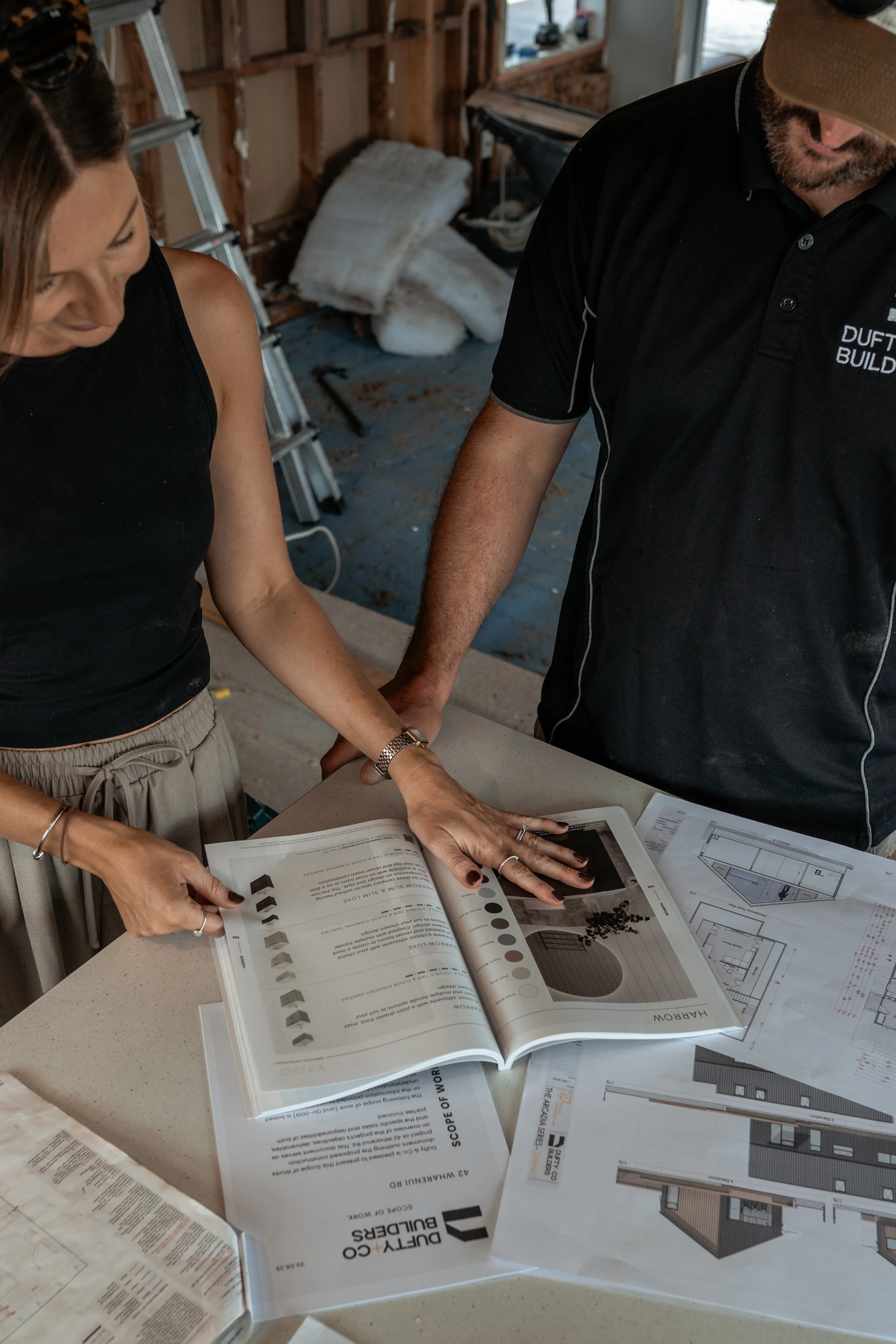 Two people looking at architectural building plans and a sample color palette on a table in a construction site or workshop.