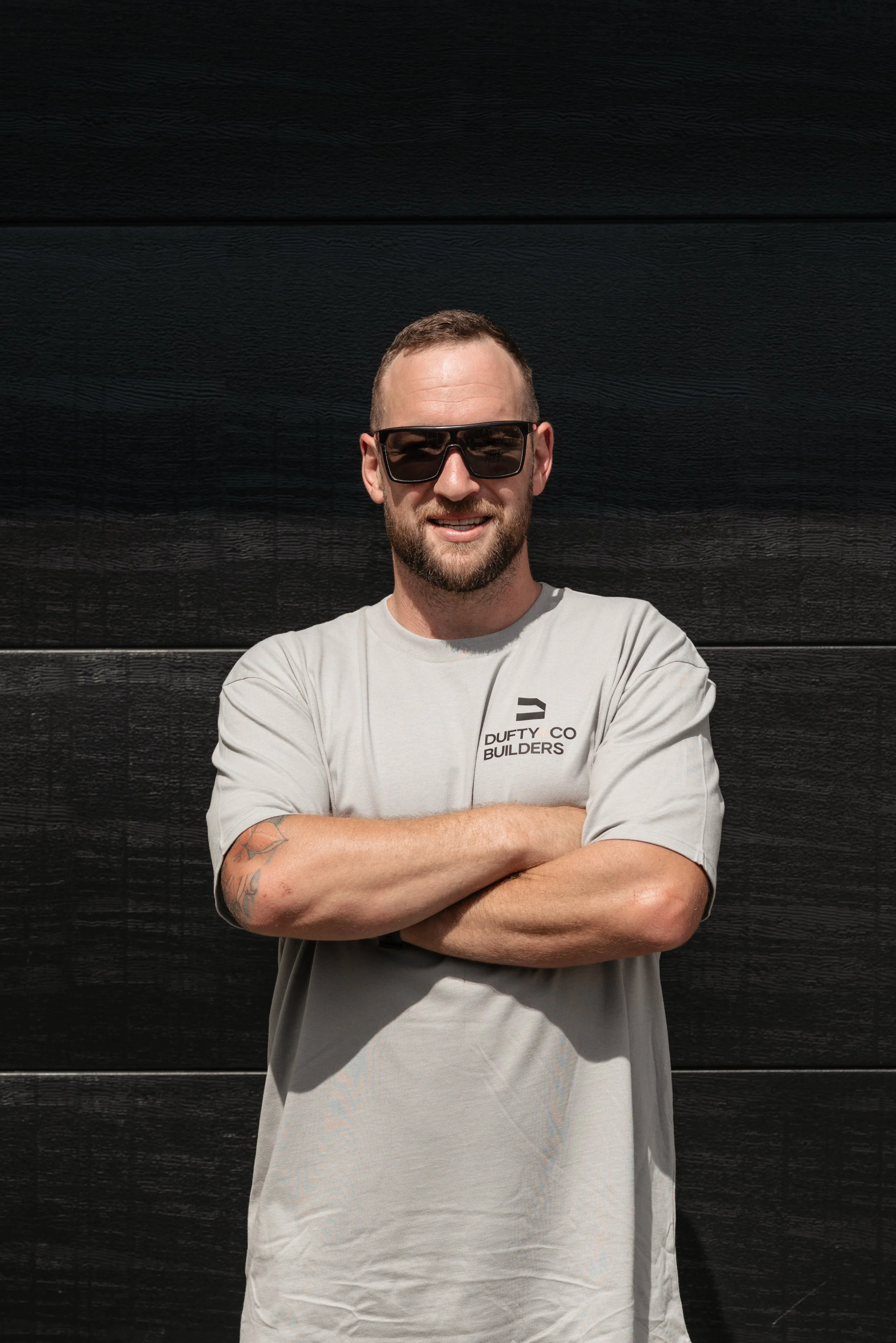 A man with a beard, wearing sunglasses and a gray T-shirt with 'Duffy Co Builders' printed on it, standing with arms crossed in front of a black wooden wall.
