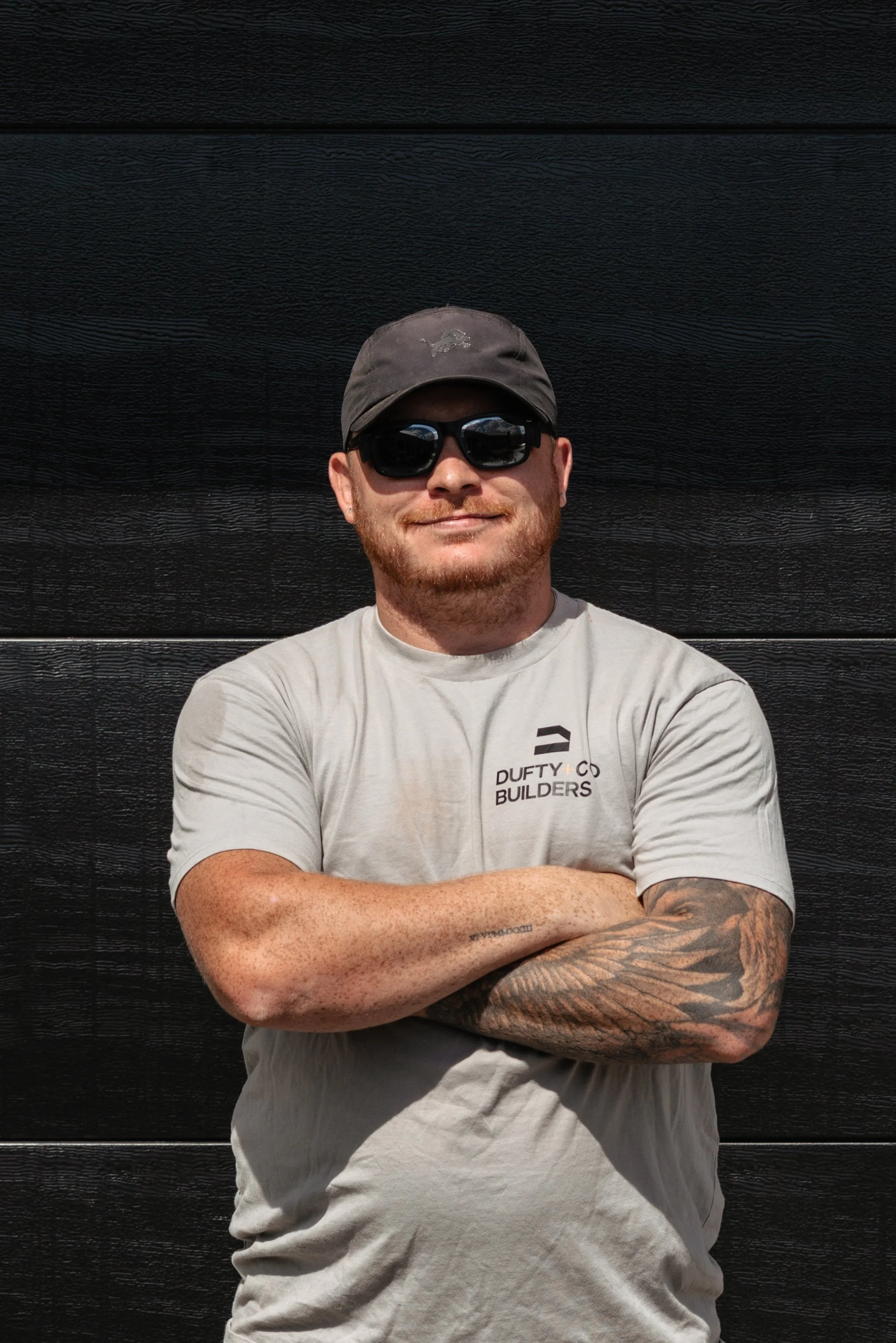 A man with a beard, sunglasses, and a black cap standing with arms crossed in front of a black wall.