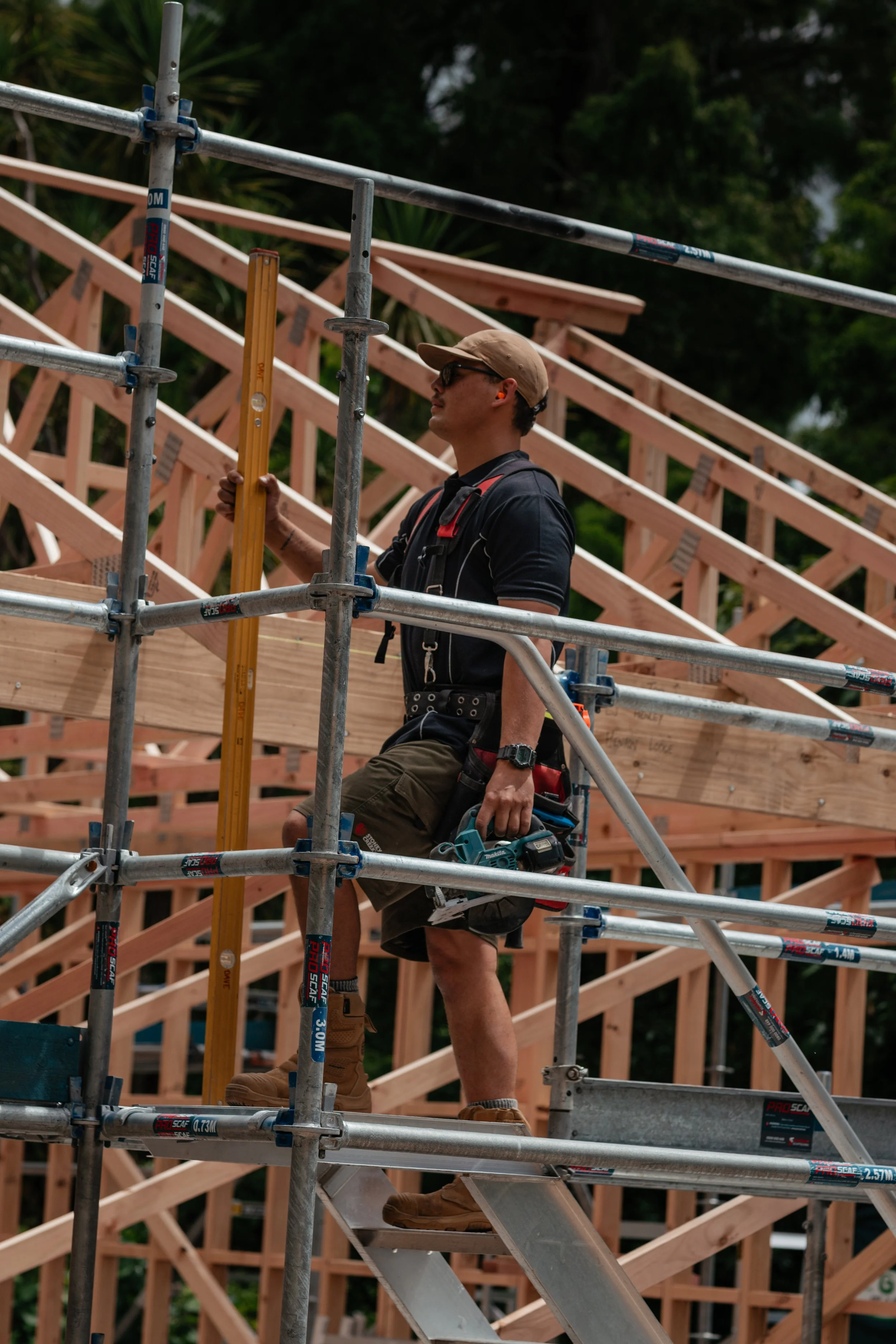 Construction worker standing on scaffolding holding a level, working on a wooden structure in an outdoor setting.