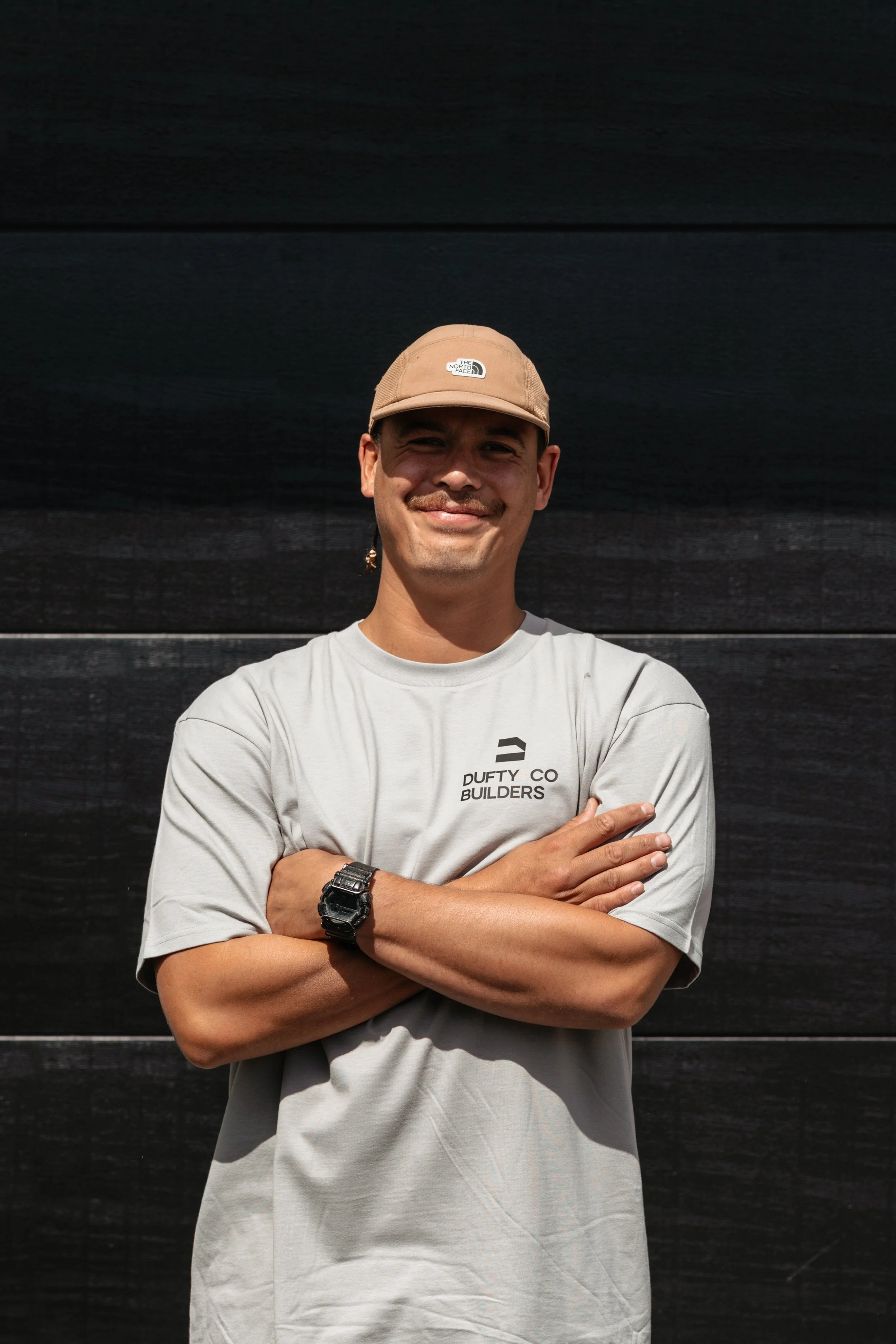 A man standing in front of a black wall, smiling with arms crossed, wearing a beige cap, a light gray t-shirt that says 'DUFTY CO BUILDERS,' and a black wristwatch.