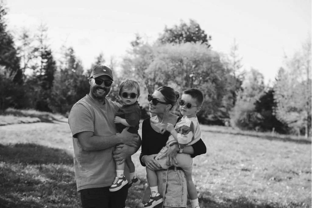 A family of four outdoors in a park, all wearing sunglasses. The man is holding a young boy, and the woman is holding another young boy. They are smiling and standing on grass with trees in the background.