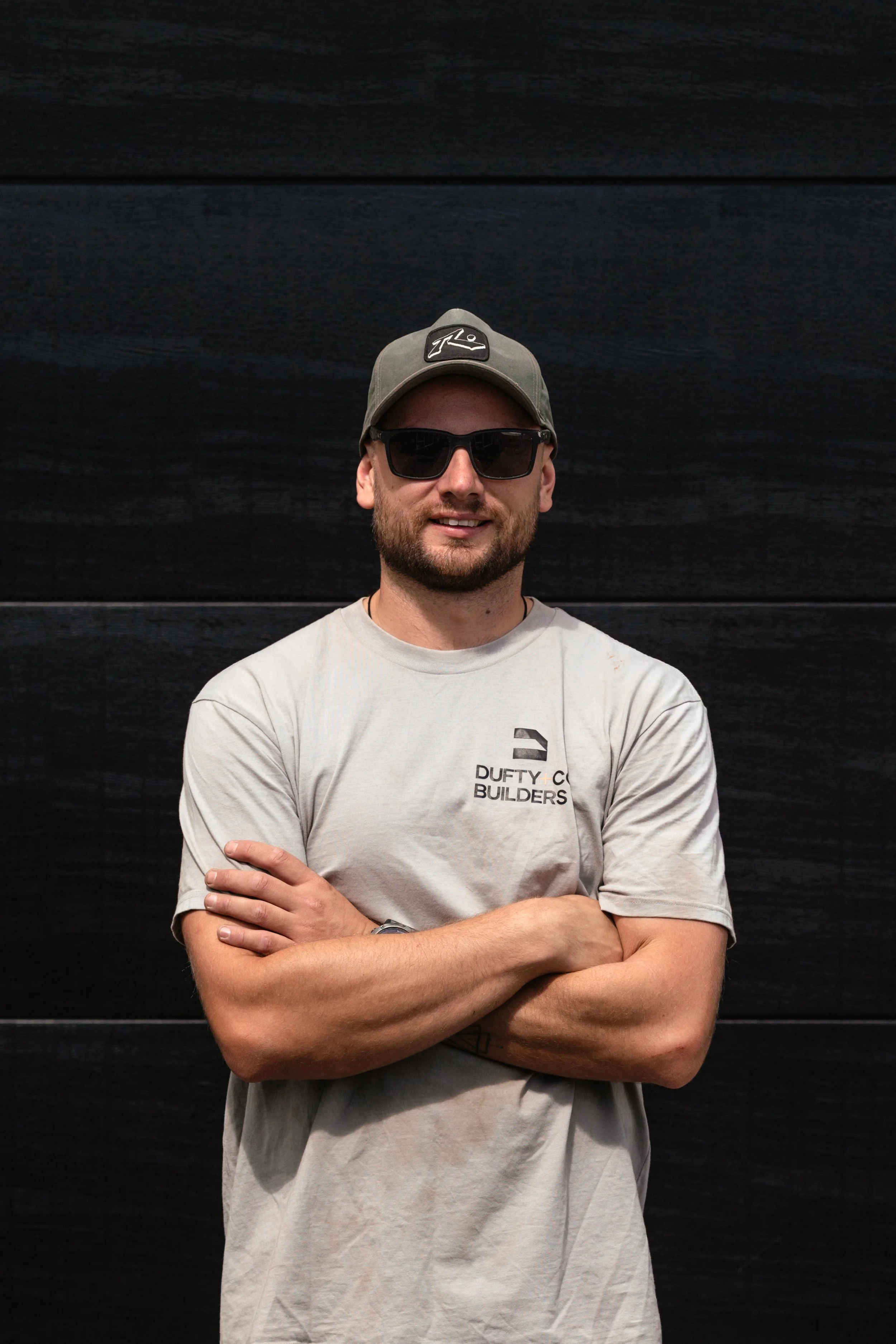 A man wearing sunglasses, a gray cap with a logo, and a light gray t-shirt with a logo and text, standing with arms crossed in front of a dark wooden wall.