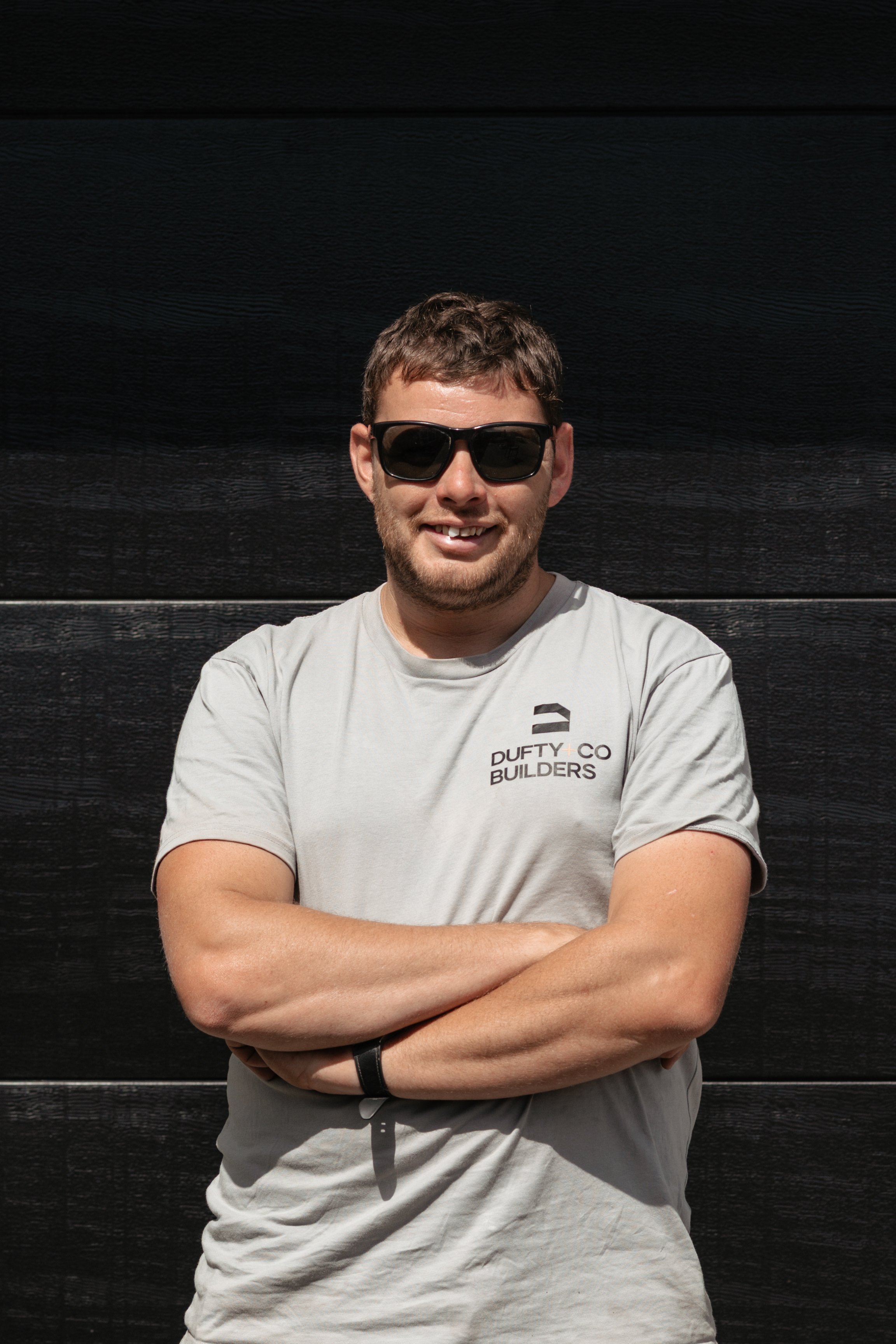 A young man with brown hair and beard, wearing black sunglasses and a light gray T-shirt with a logo that says 'Duffy Co Builders', stands with his arms crossed in front of a dark wooden wall, smiling at the camera.