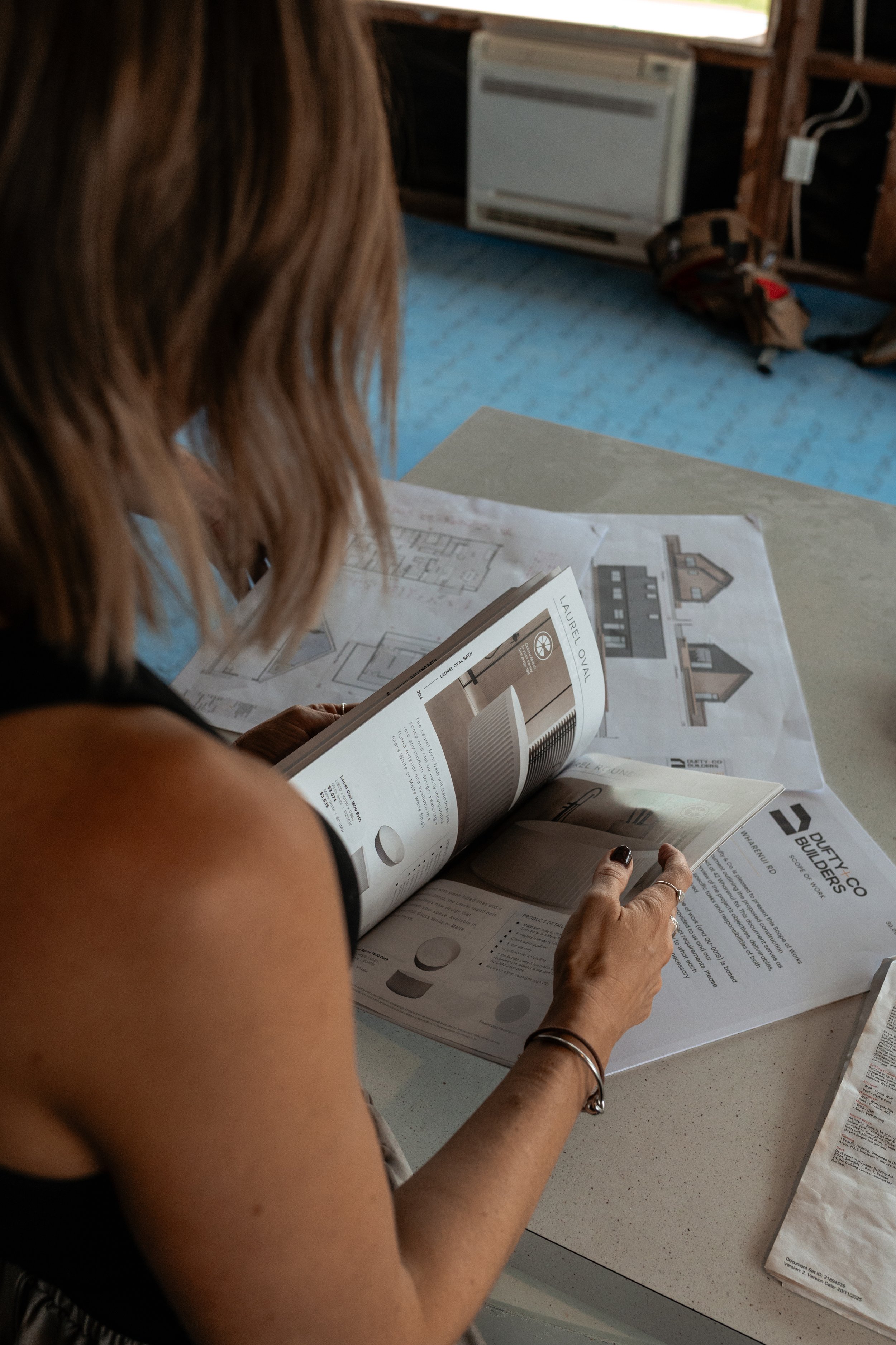 A woman with light brown hair looking at an open catalog on a table, with architectural plans and a brochure about 'Laura Ova' blueprints and the 'Identity Co Builders' logo nearby.