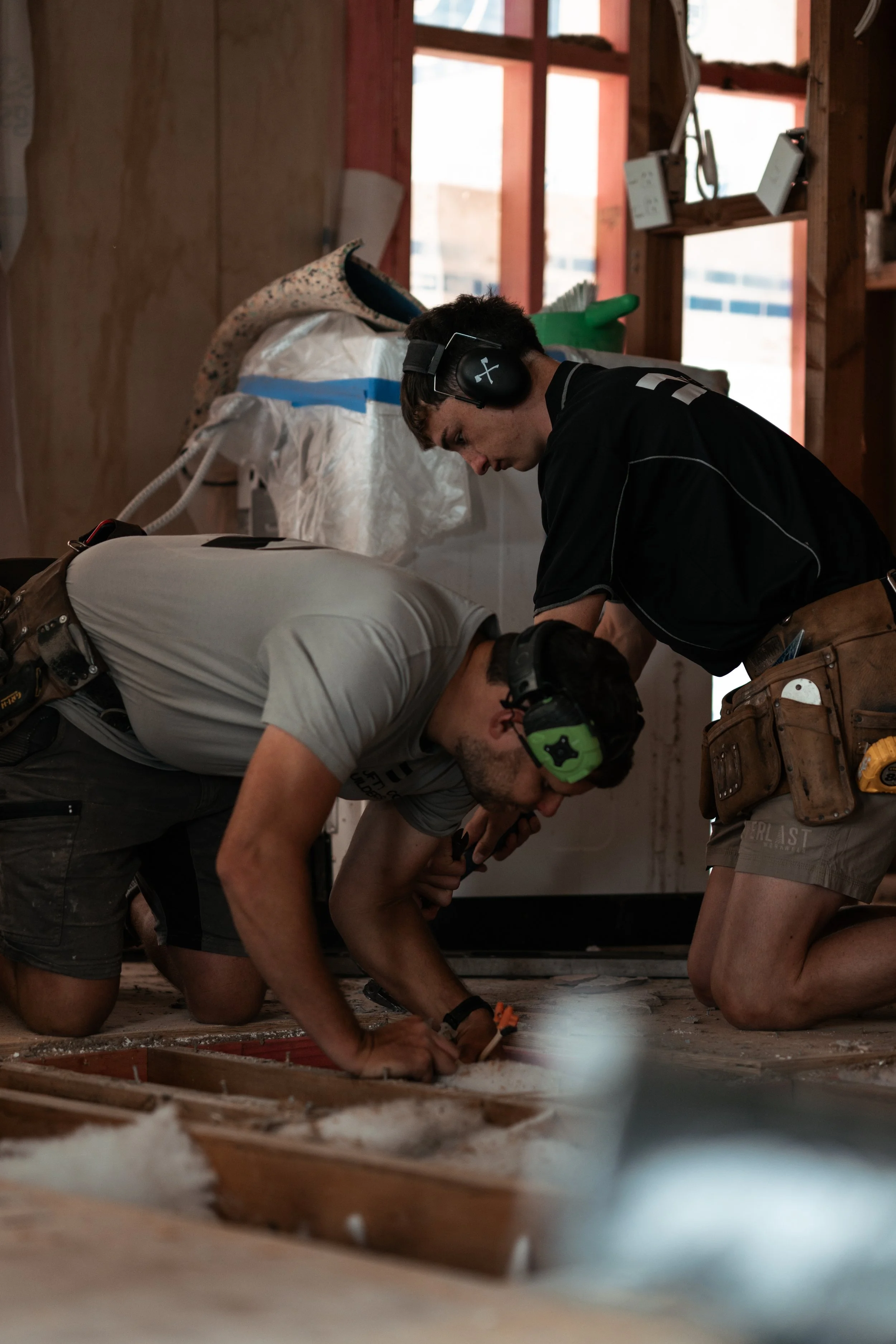 Two construction workers kneel on the floor working on a floorboard in a partially built room, with construction tools and equipment nearby.
