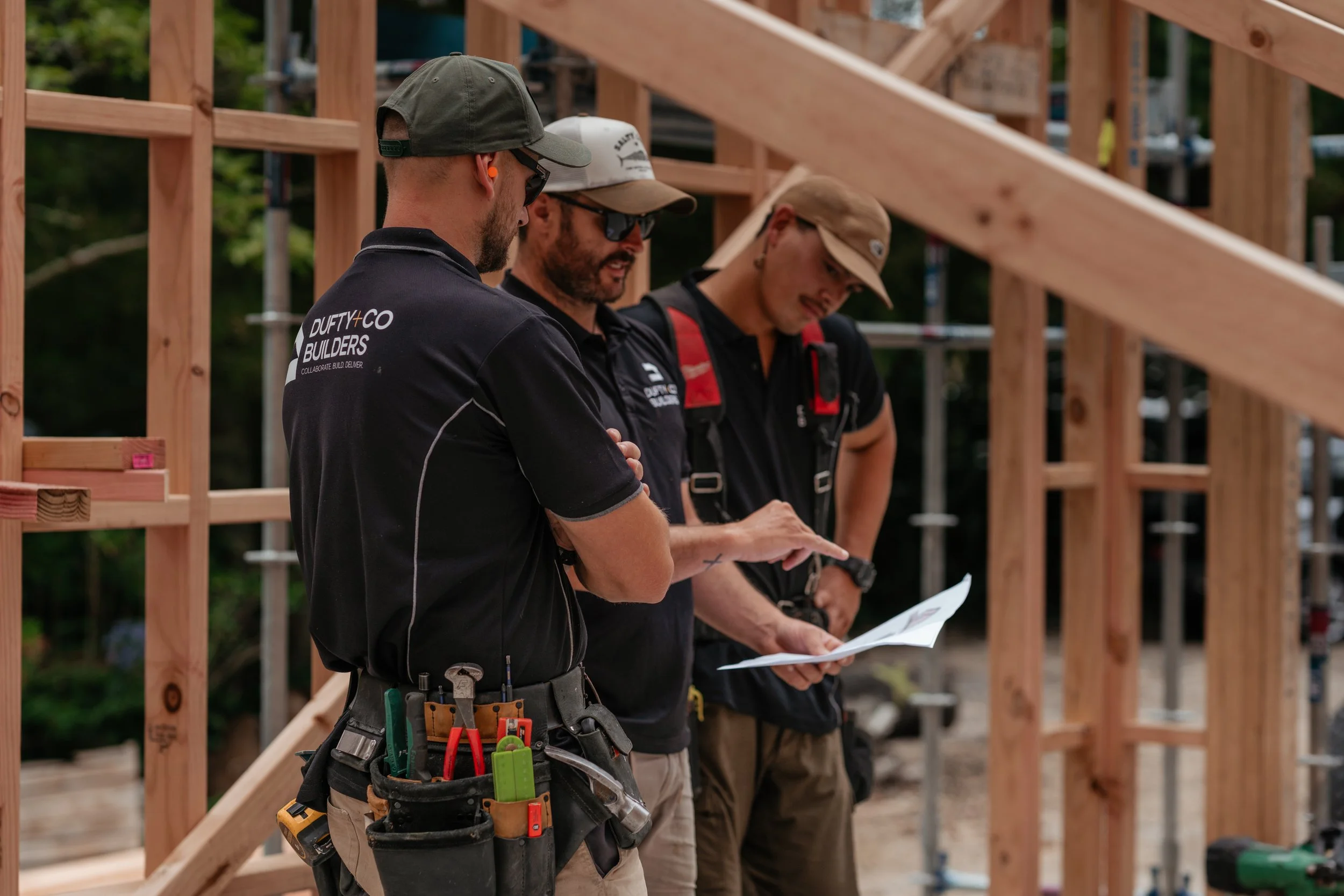 Three construction workers reviewing plans at a building site with a wooden frame in the background.