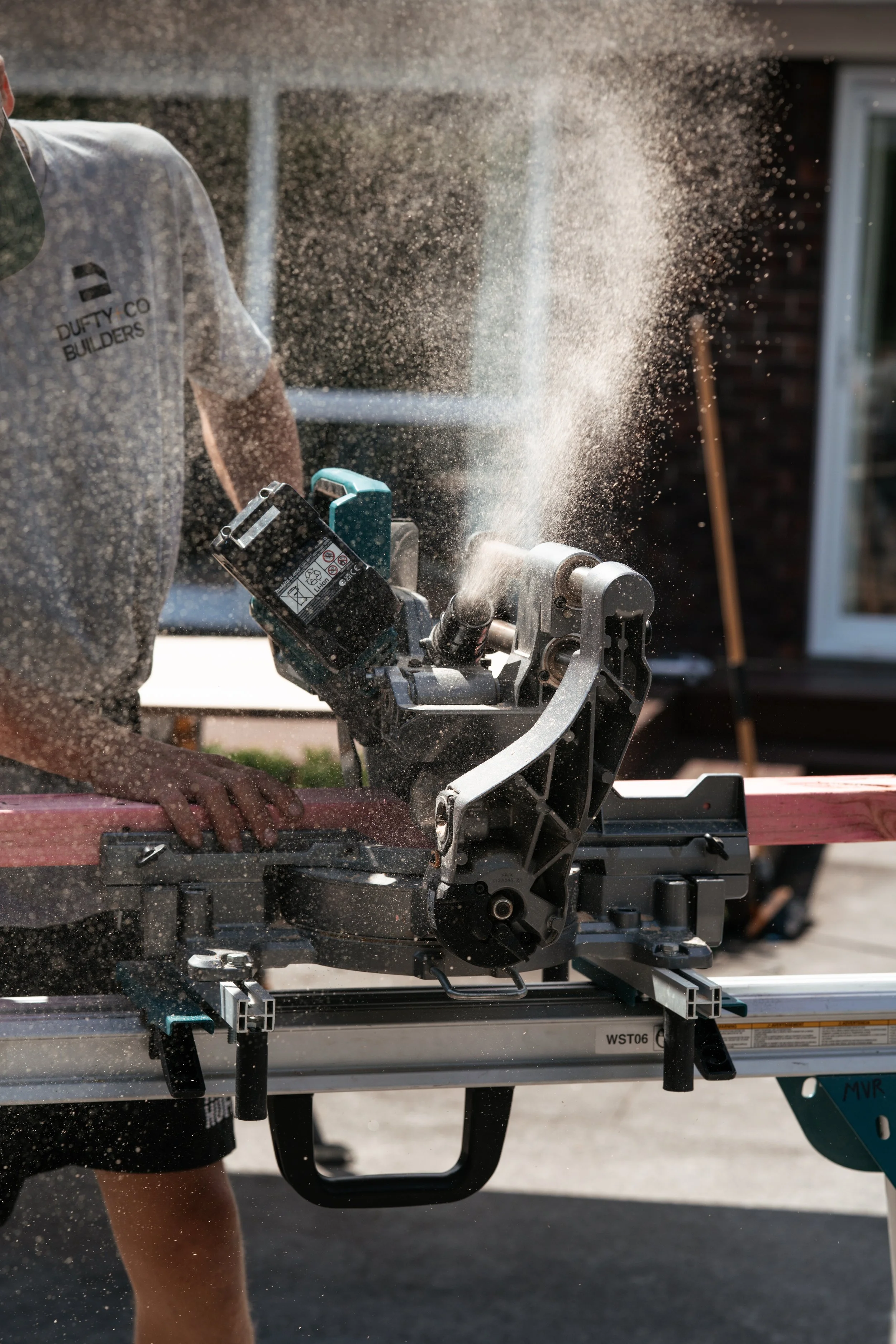 Person cutting wood with a powered miter saw outdoors, with sawdust flying in the air.