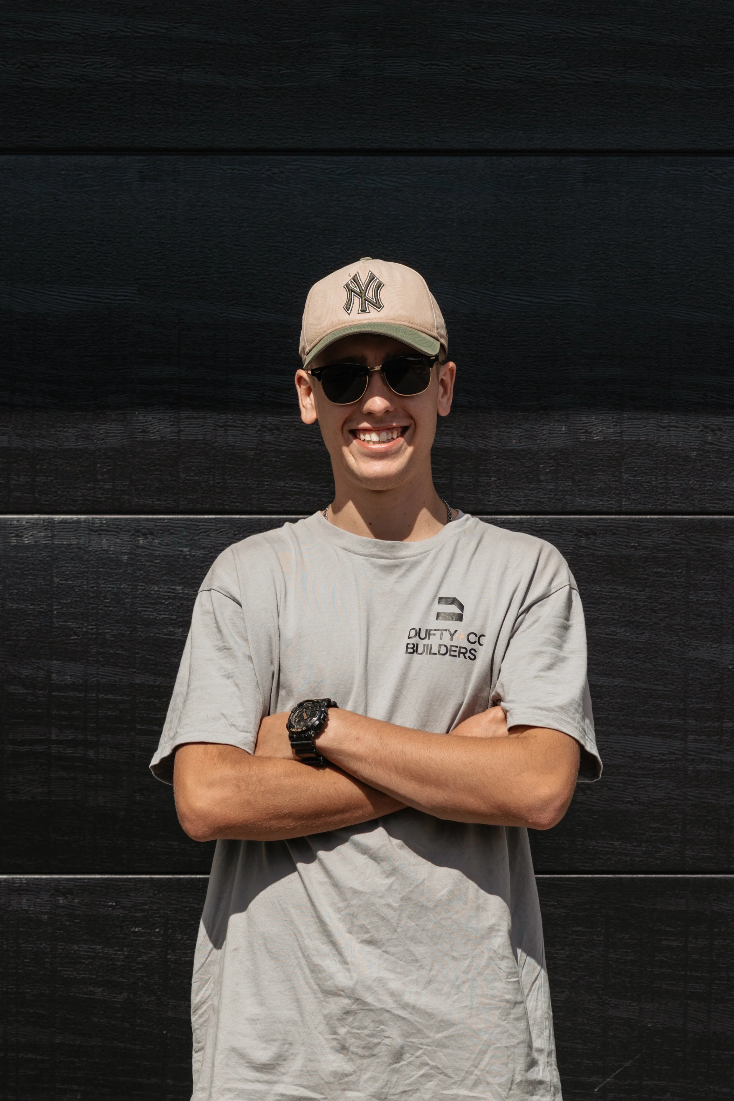 A young man standing with crossed arms in front of a black wooden wall. He is wearing a beige baseball cap, sunglasses, a light gray T-shirt with a logo, and a black wristwatch. He is smiling.