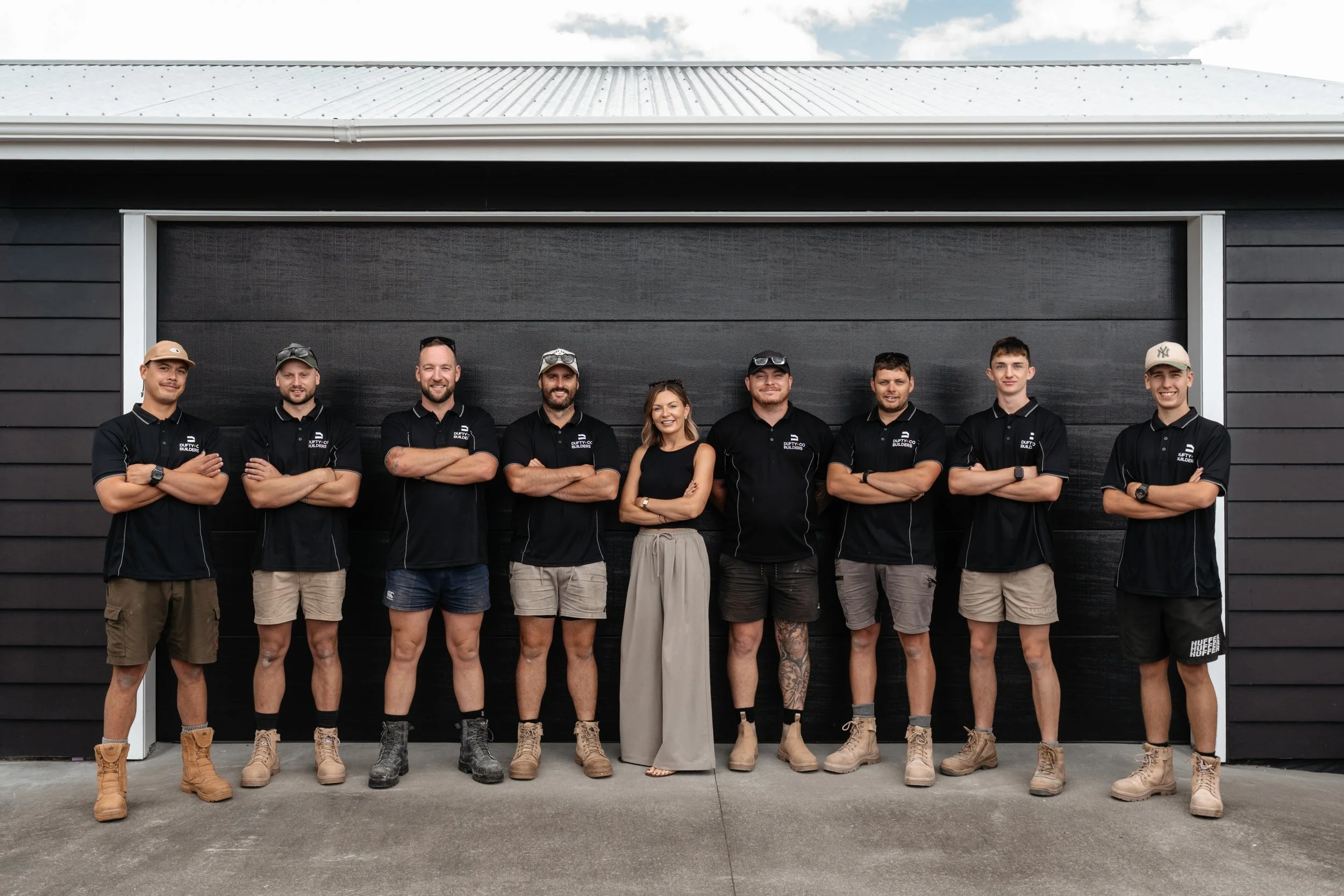 Group of nine people standing in front of a black garage door, posing for a photo. They are wearing black shirts with logos, shorts or pants, and work boots. One woman is in the center, wearing a black sleeveless top and beige wide-legged pants. The group appears to be outdoors on a concrete driveway.