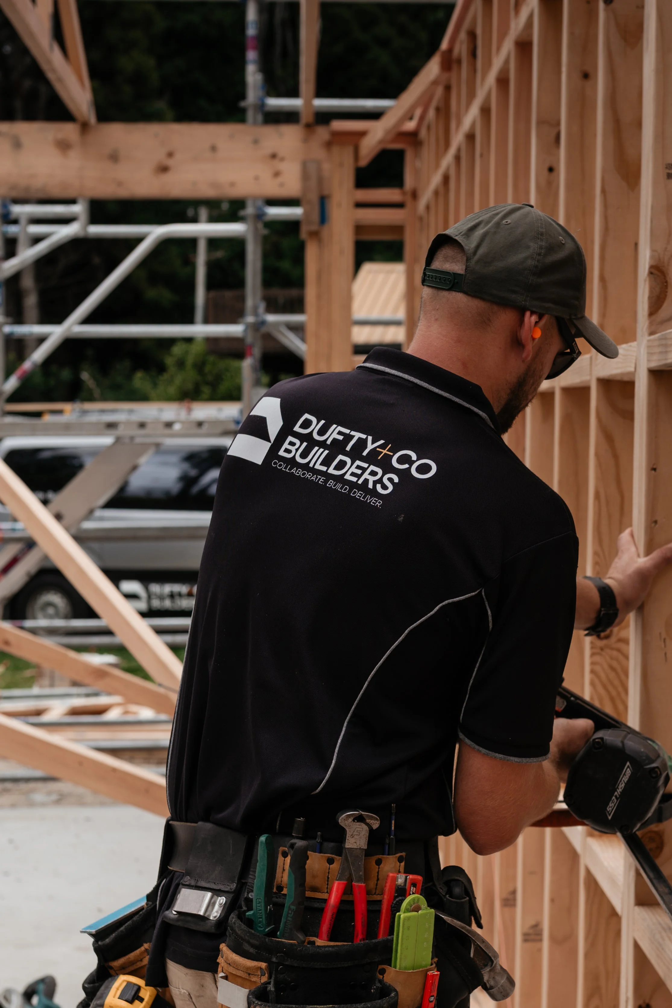 A construction worker building a wooden structure on a construction site, wearing a black shirt, cap, and tool belt with various tools.