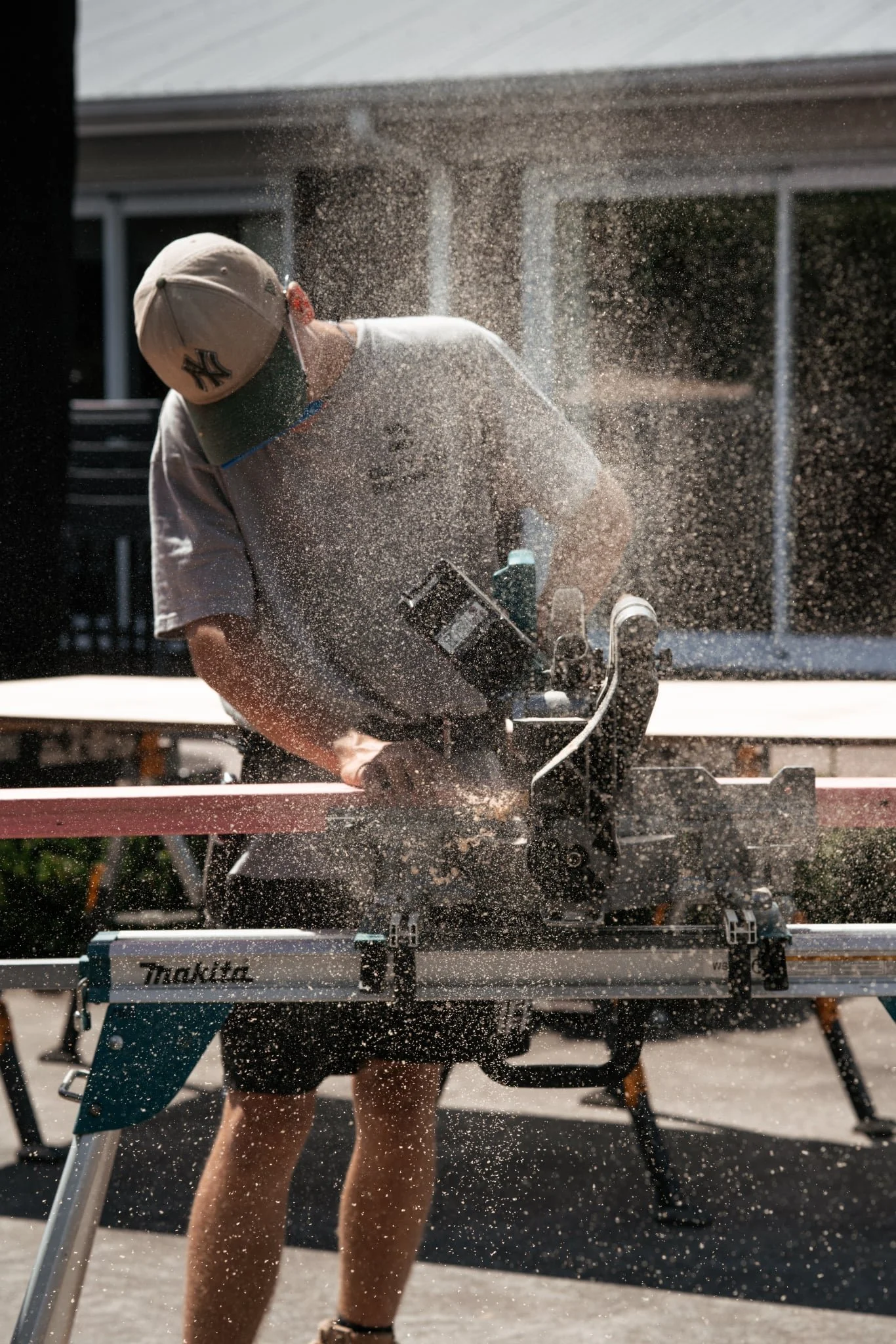 Person wearing a baseball cap working with a power saw, sawdust flying in the air, outdoor setting.
