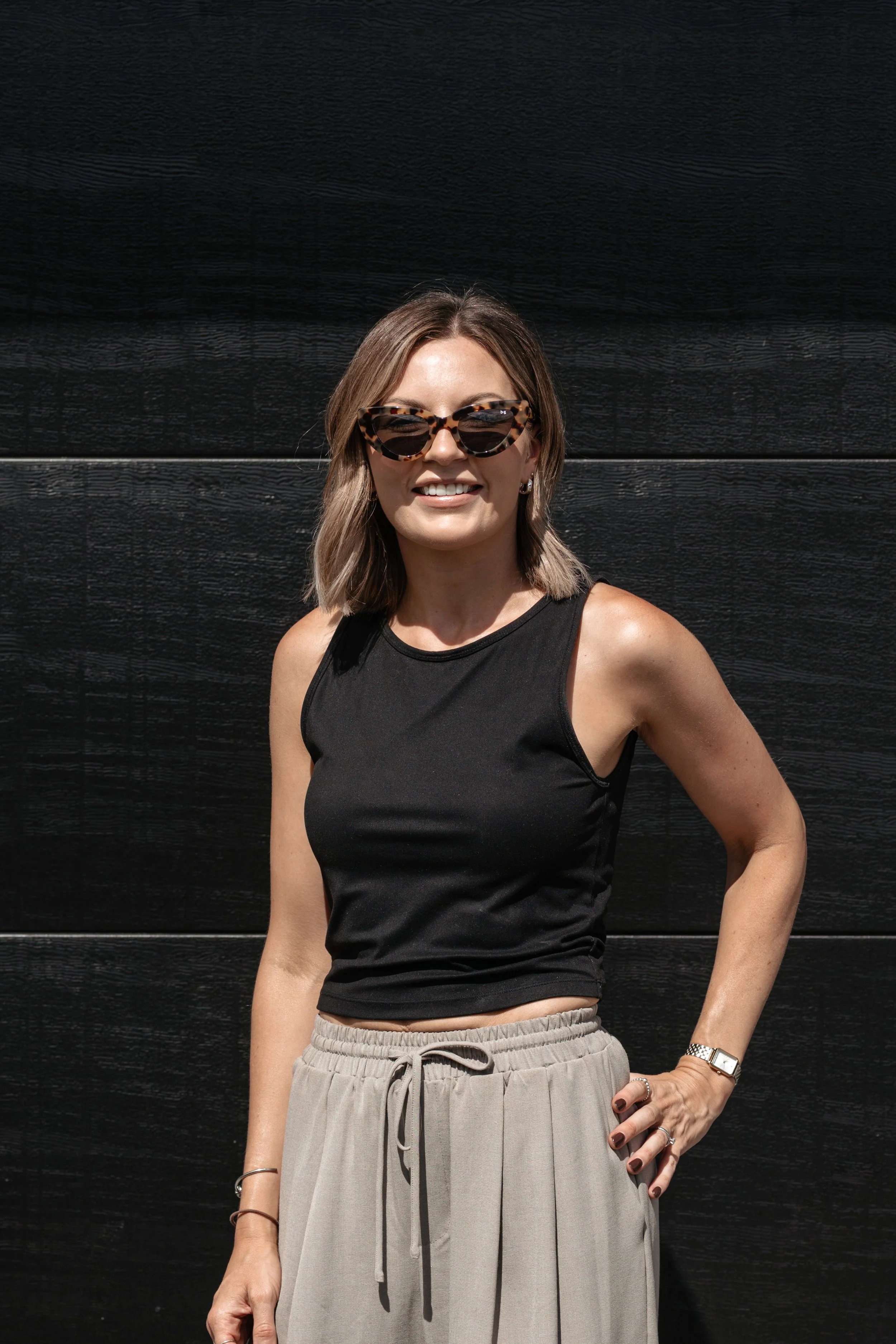 Woman with shoulder-length hair wearing sunglasses, a black sleeveless top, and beige pants standing against a black wooden wall.
