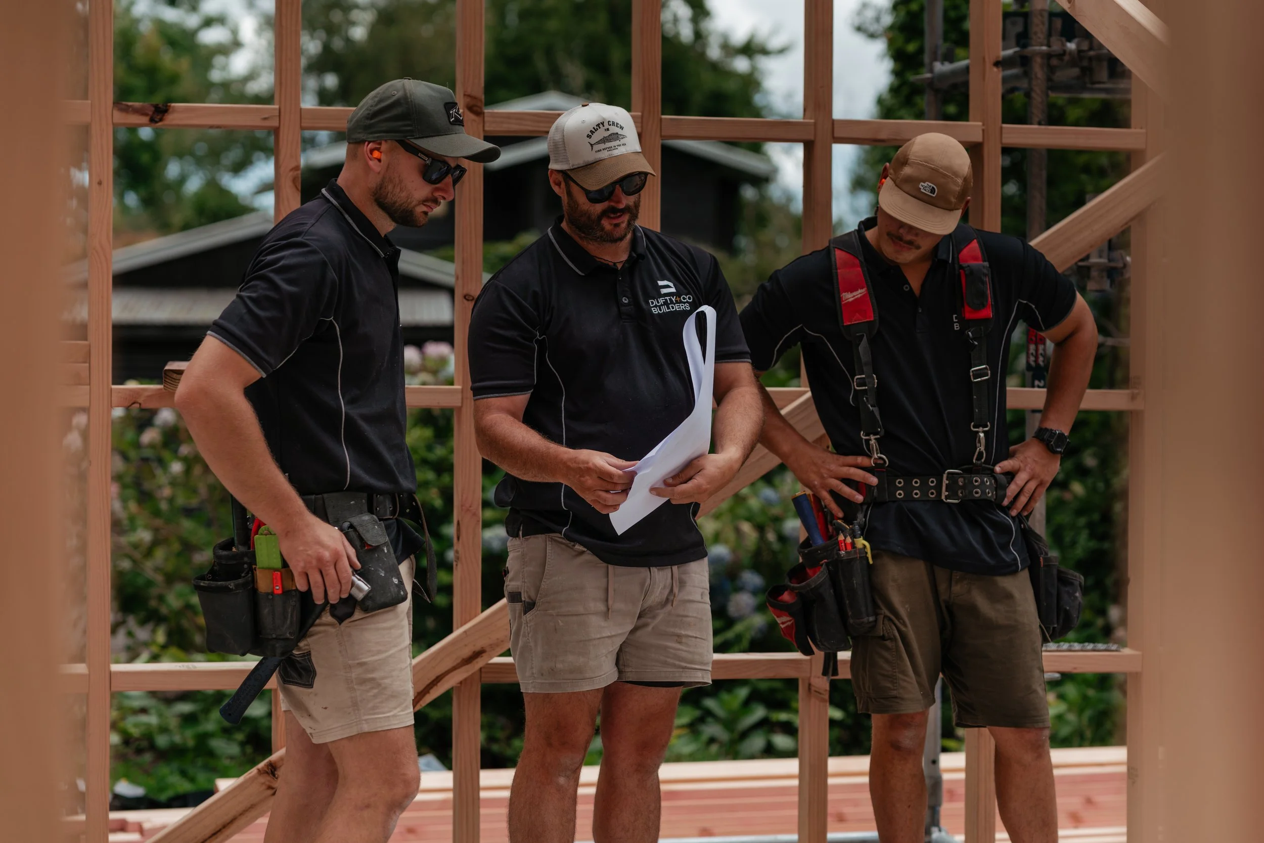 Three construction workers reviewing plans on a wooden construction site frame.