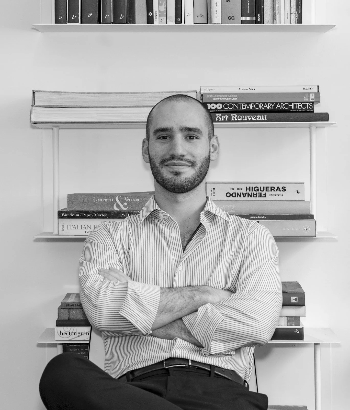 Retrato en blanco y negro de un hombre con barba y cabeza calva, vestido con una camisa de rayas, con los brazos cruzados, sentando frente a una estantería con libros.