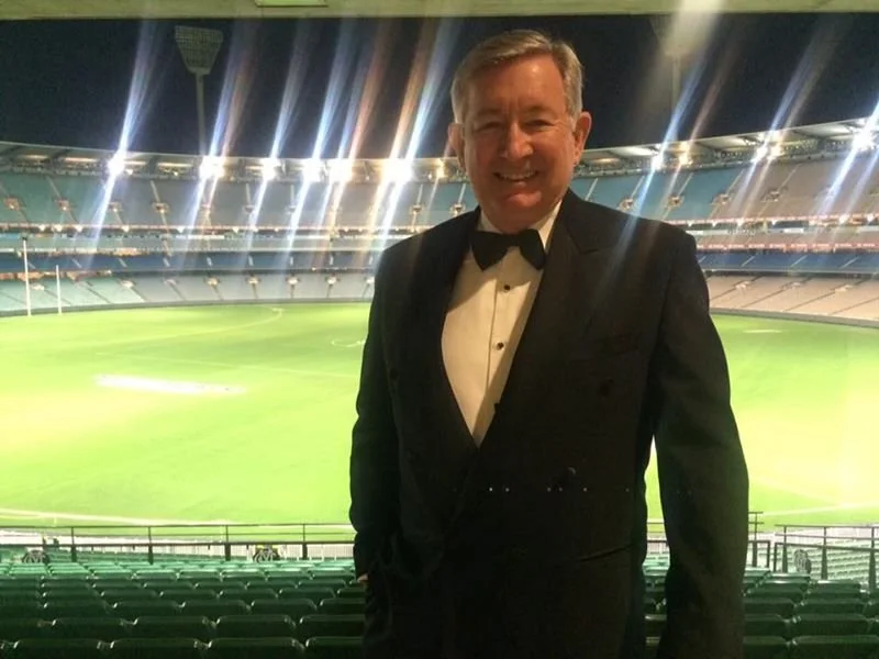 A man in a tuxedo standing in a stadium, smiling at the camera, with an empty field and seating in the background.