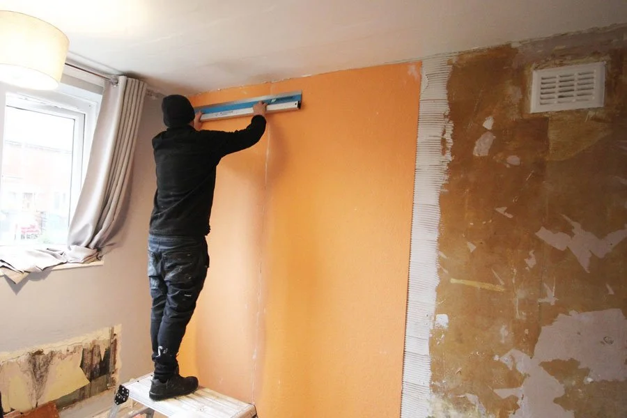A person standing on a ladder and applying orange wall paint in a room that is undergoing renovation.