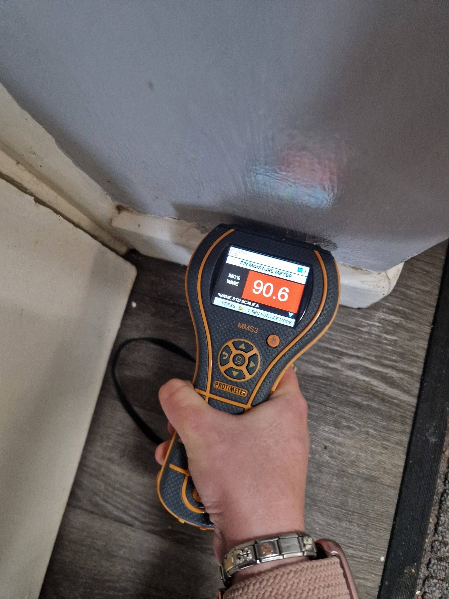 A person holding a digital moisture meter against a wall, displaying a moisture level of 90.6.