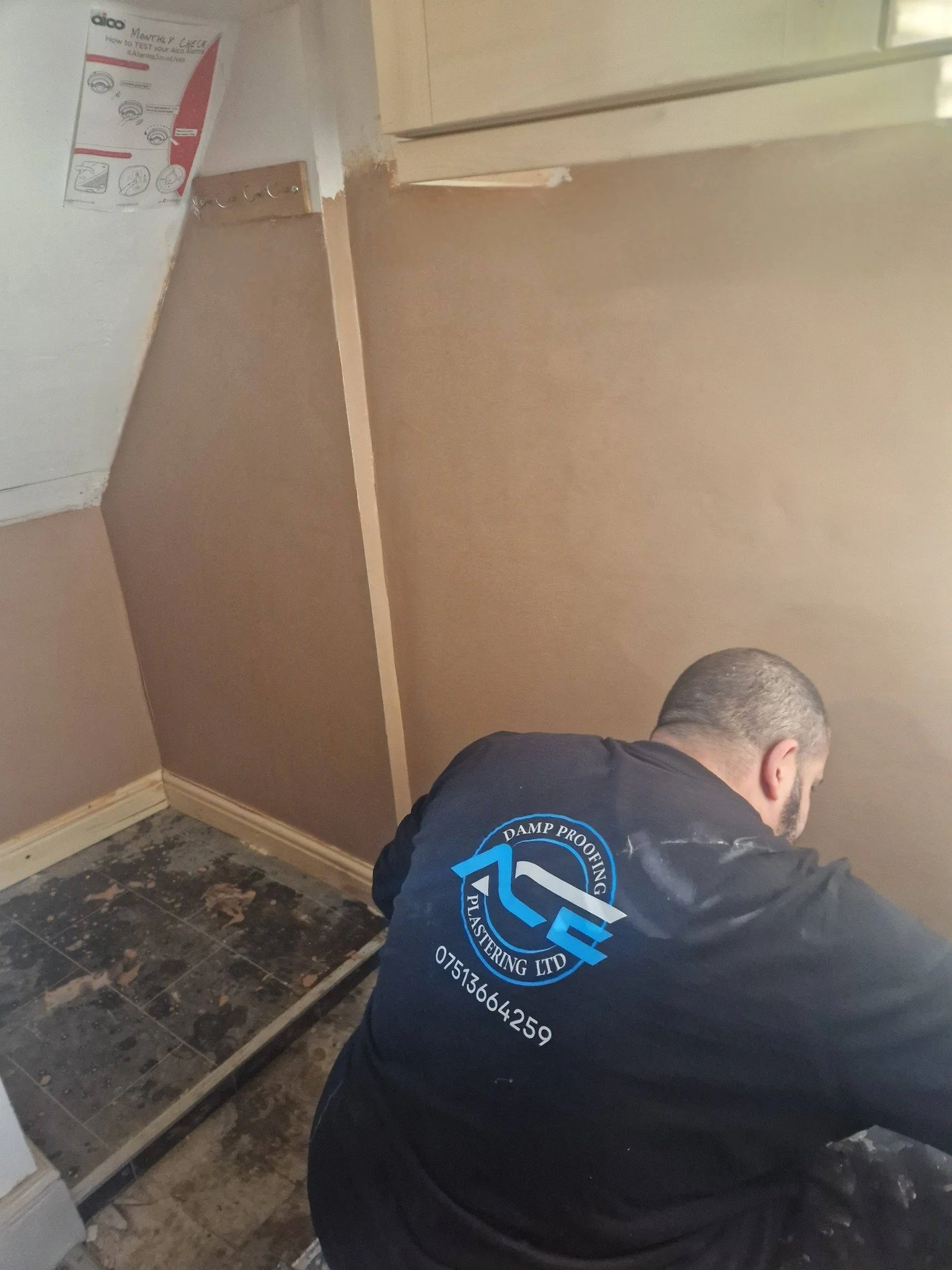 A man in a black jacket with a logo and phone number on the back is working on a wall, possibly drywall or painting, in a room with a partially removed or damaged floor. The room appears to be under renovation.