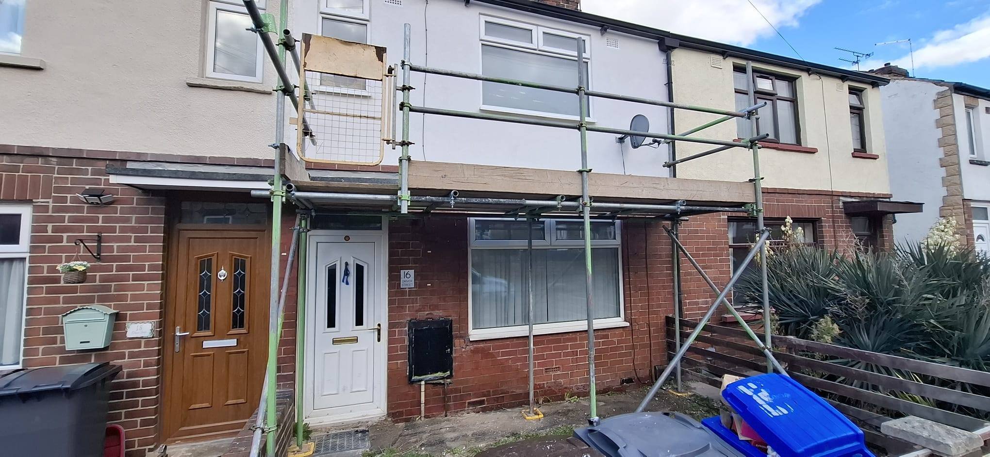 House with scaffolding and construction work on the front facade, two doors, a window, and construction materials in the yard.
