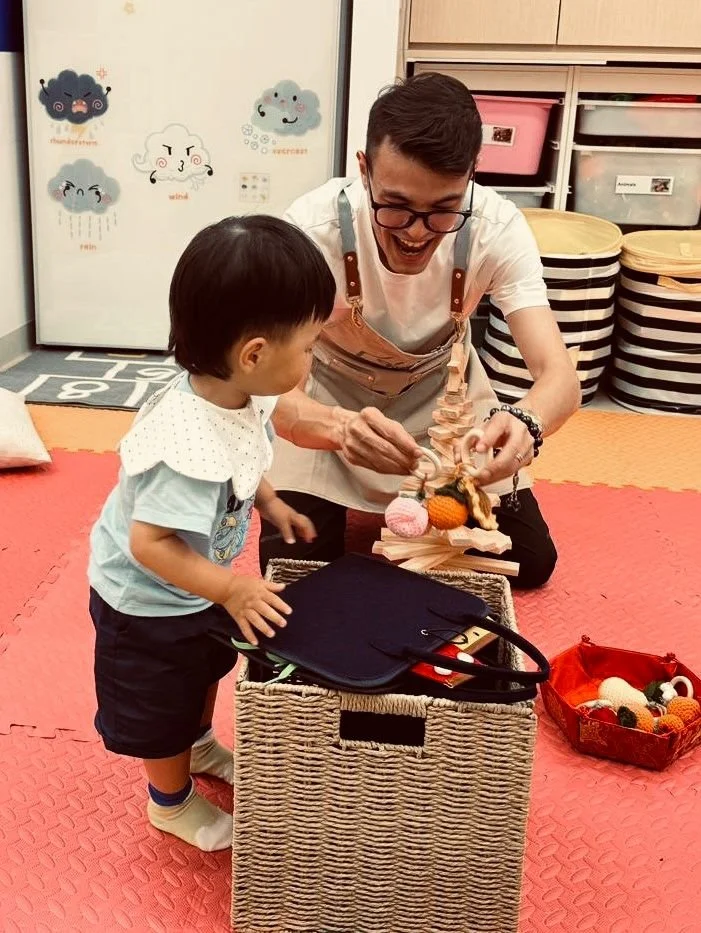 An adult and a young child playing together in a colorful indoor space, with various toys and storage bins around. The adult is smiling and holding a puppet, engaging the child who is looking at the puppet with interest.