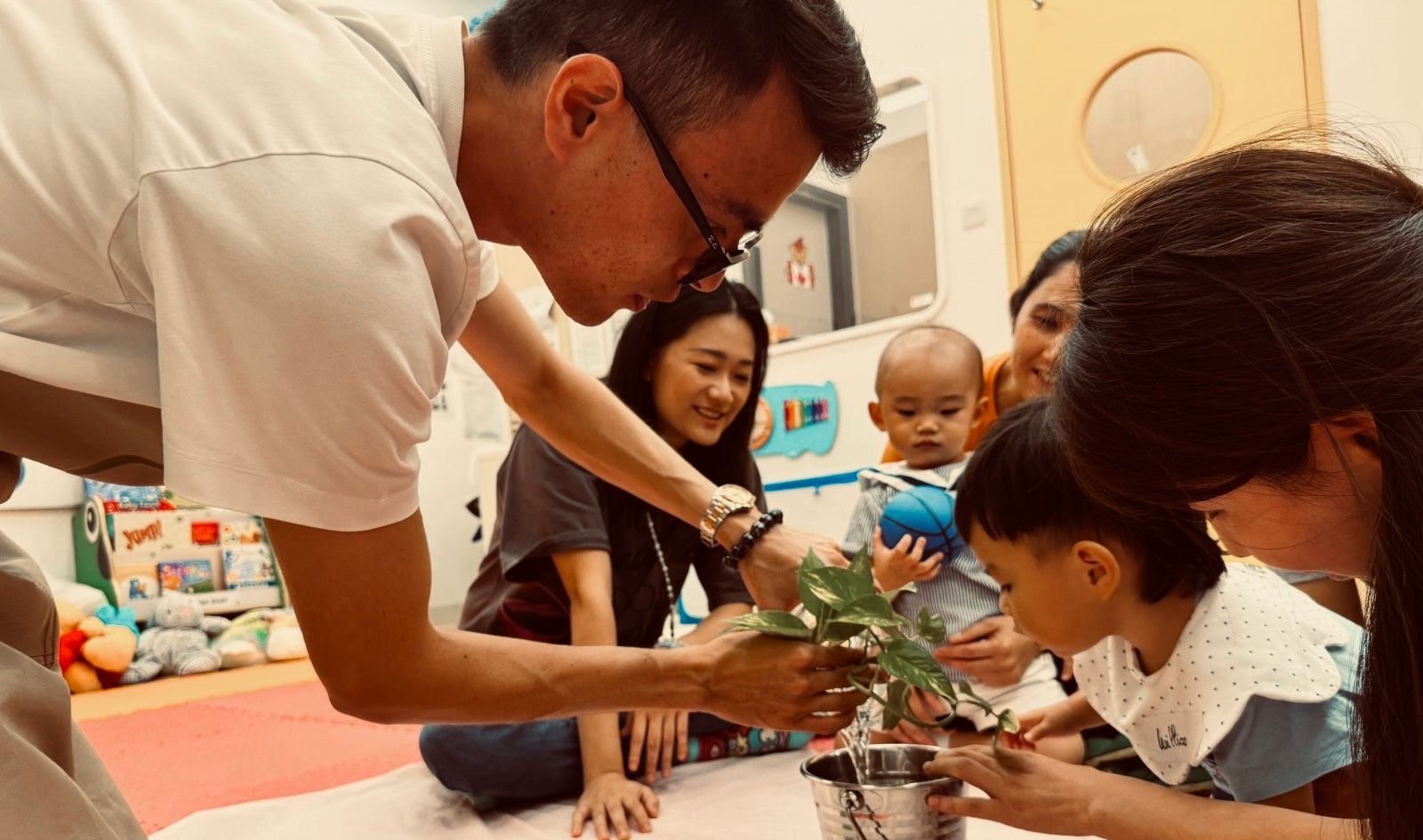 A man in a white shirt plants a small leafy plant in a metal pot with children and women gathered around watching and smiling, in a brightly lit room.