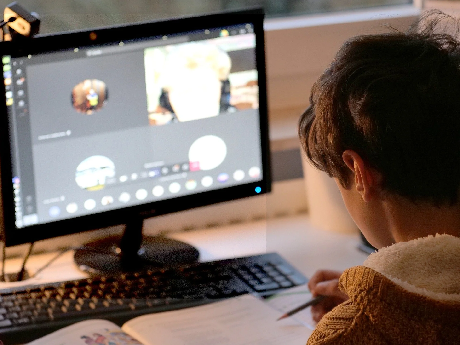A child with brown hair sitting at a desk, using a desktop computer with a large monitor displaying a photo editing or drawing software. The child appears to be working on a project, with a notebook and pencil on the desk.