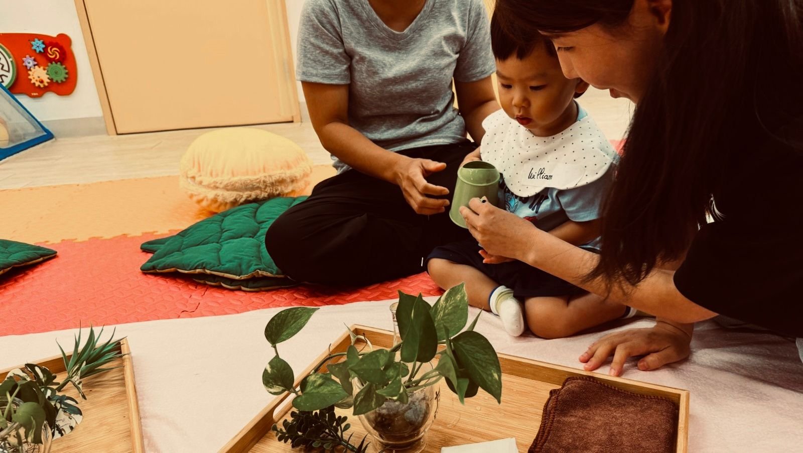 Young child being shown a cup by two adults in a cozy, decorated room with plants, cushions, and educational decor.