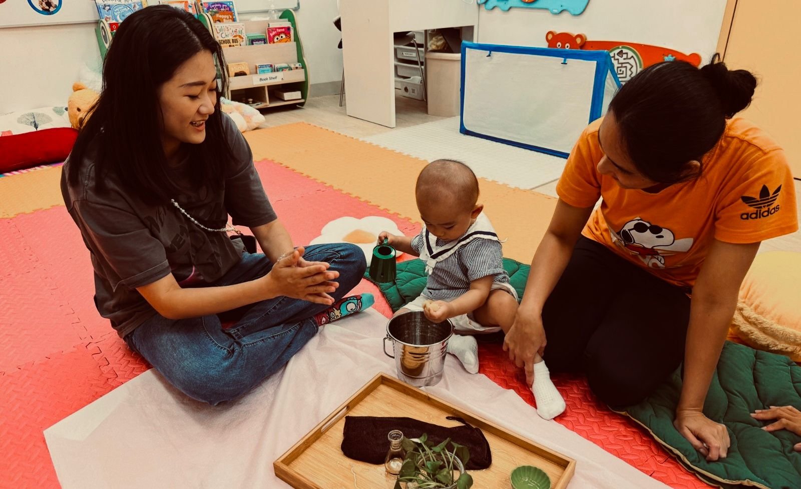A young boy is sitting on a pink blanket with two women, one in a black shirt and the other in an orange shirt. The boy is holding a green watering can over a small metal bucket, and the women are smiling and watching him. The setting appears to be a