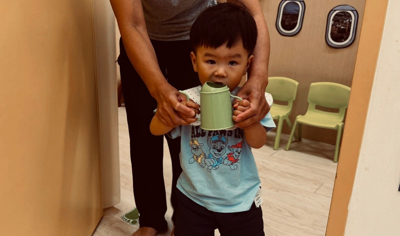 A young boy holding a green watering can, with an adult standing behind him, in a room with beige walls and green chairs.