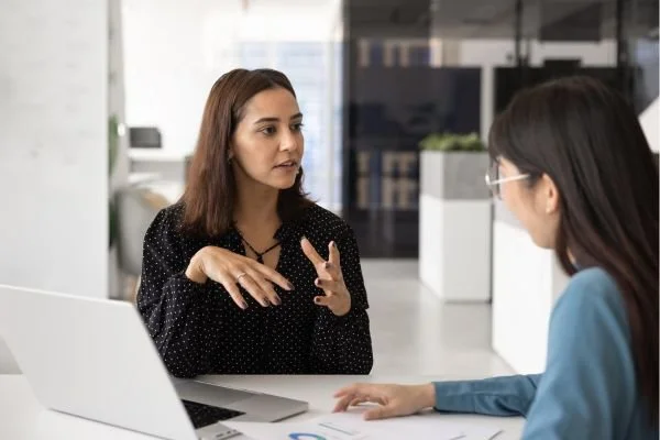 A woman in a black patterned blouse gestures with her hands while speaking with a colleague across a desk in a modern office