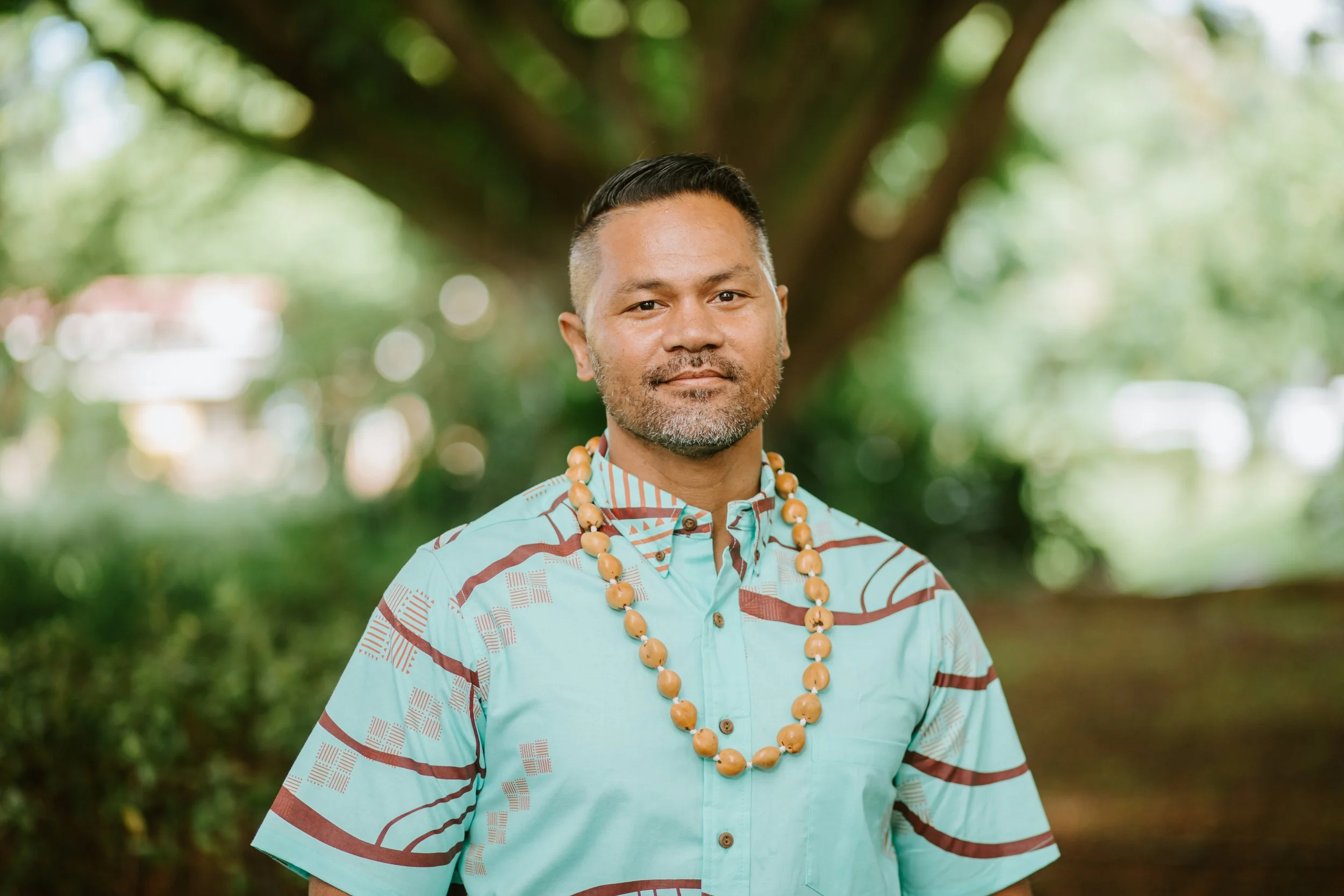 Jr in a patterned turquoise shirt wearing a kukui nut lei, standing outdoors with greenery in the background.