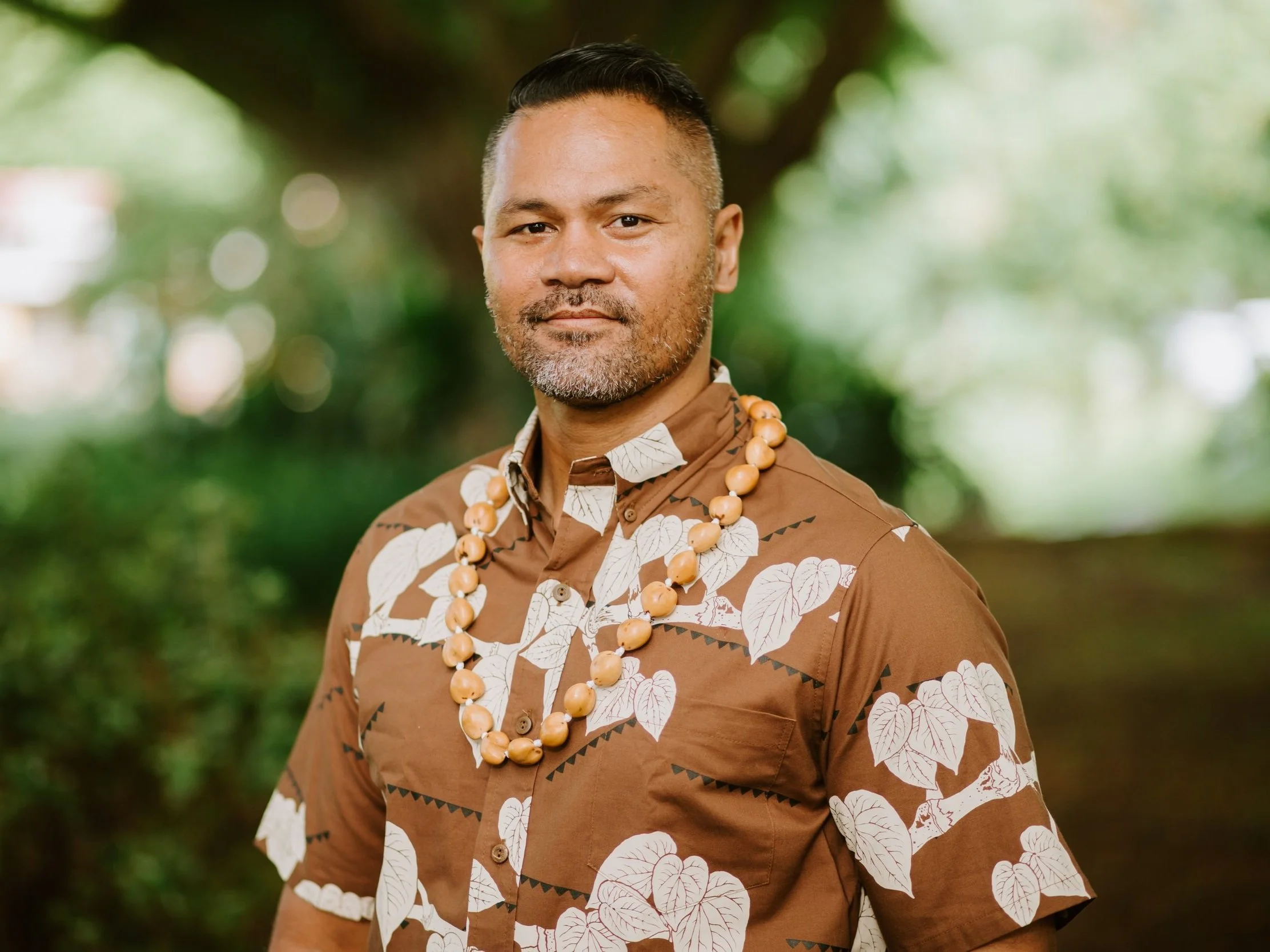 Jr in a brown Hawaiian shirt with leaf patterns, accessorized with a kukui nut lei, standing outdoors with a blurry, natural background.